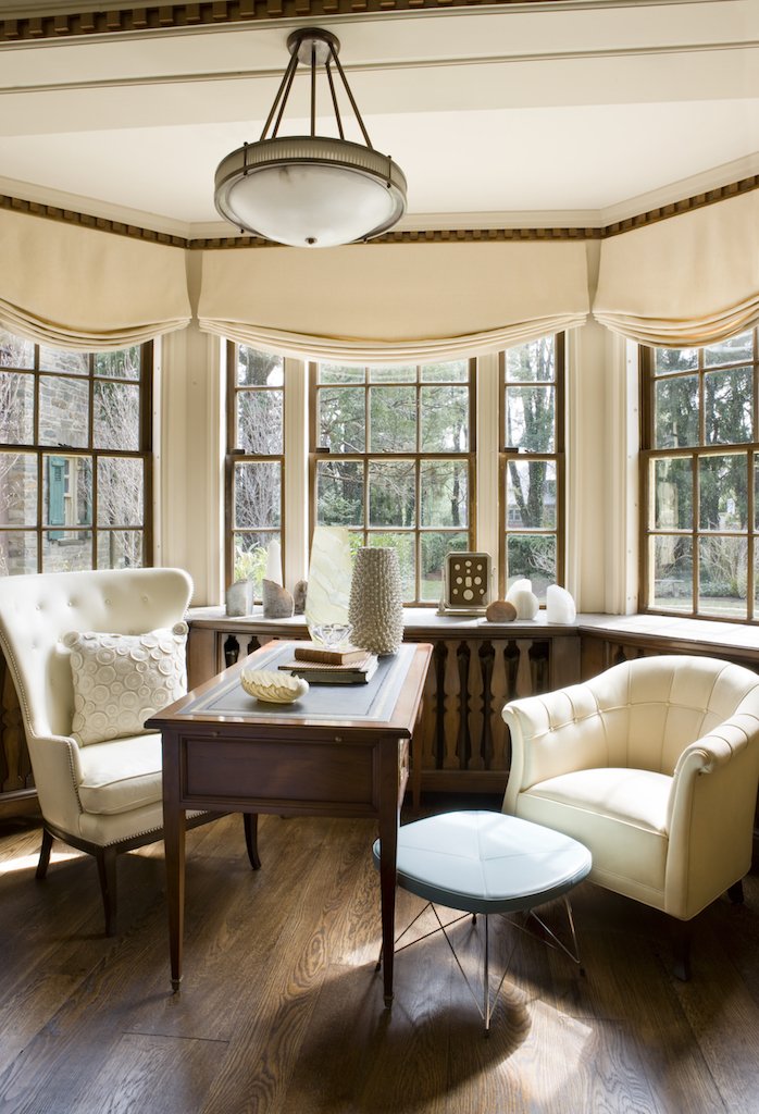 Cozy corner with a wooden desk, two cream-colored armchairs, decorative vases, and a large window with a view of trees outside.