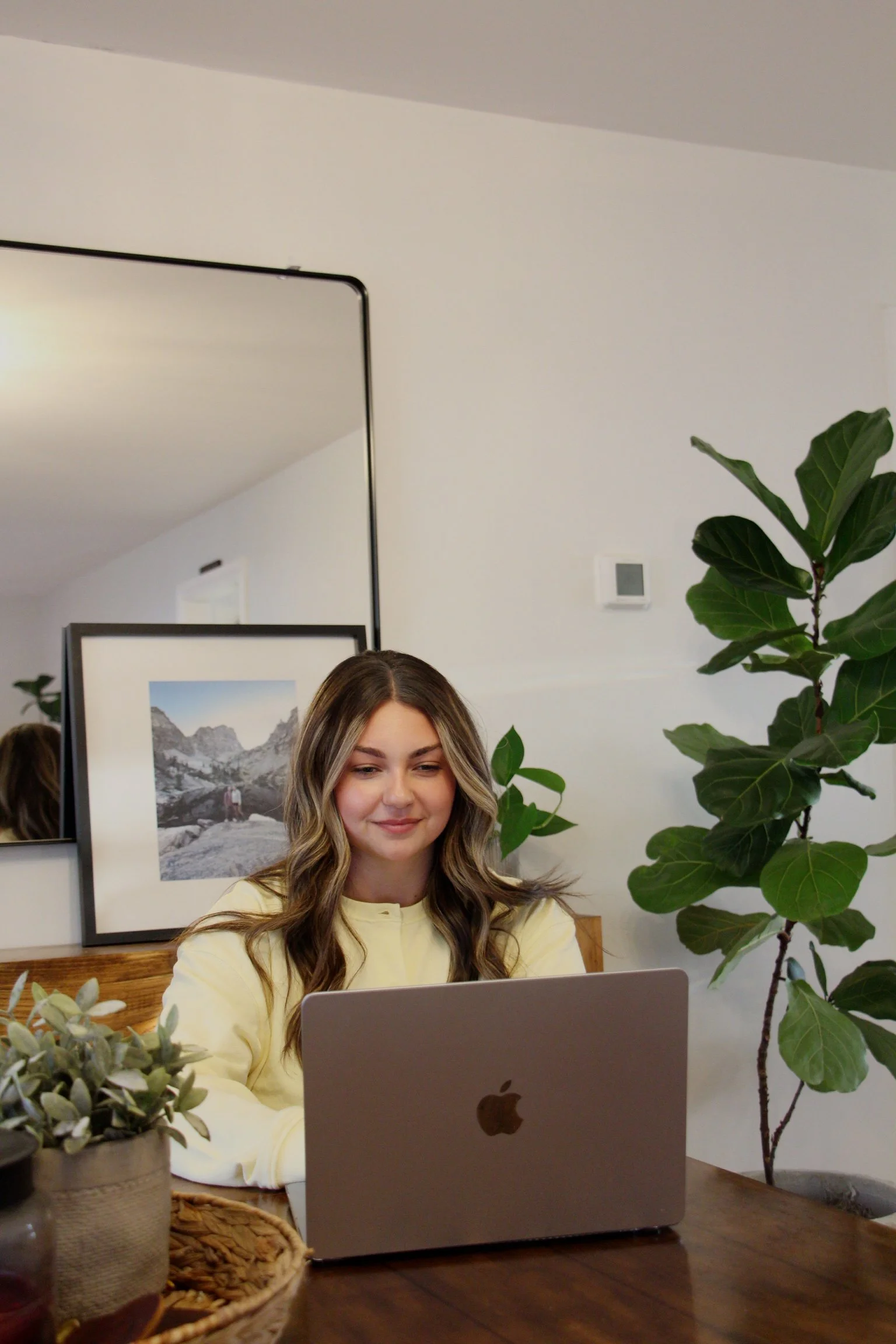 A woman with wavy, shoulder-length hair sitting at a wooden table with an open MacBook in front of her. She is wearing a yellow top and is smiling slightly while looking at the laptop screen. There are green indoor plants on the table and in the background. Behind her, there's a framed black and white landscape photograph and a white wall with a thermostat and a ceiling light. The setting appears to be a cozy, modern home or office space.