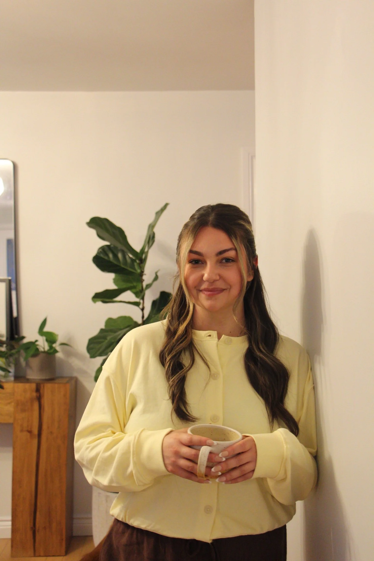 A young woman with long wavy hair, wearing a yellow button-up shirt, is standing indoors, holding a beige mug with both hands. Behind her are potted plants and a wooden cabinet, with a plain wall and a radiator to her right.