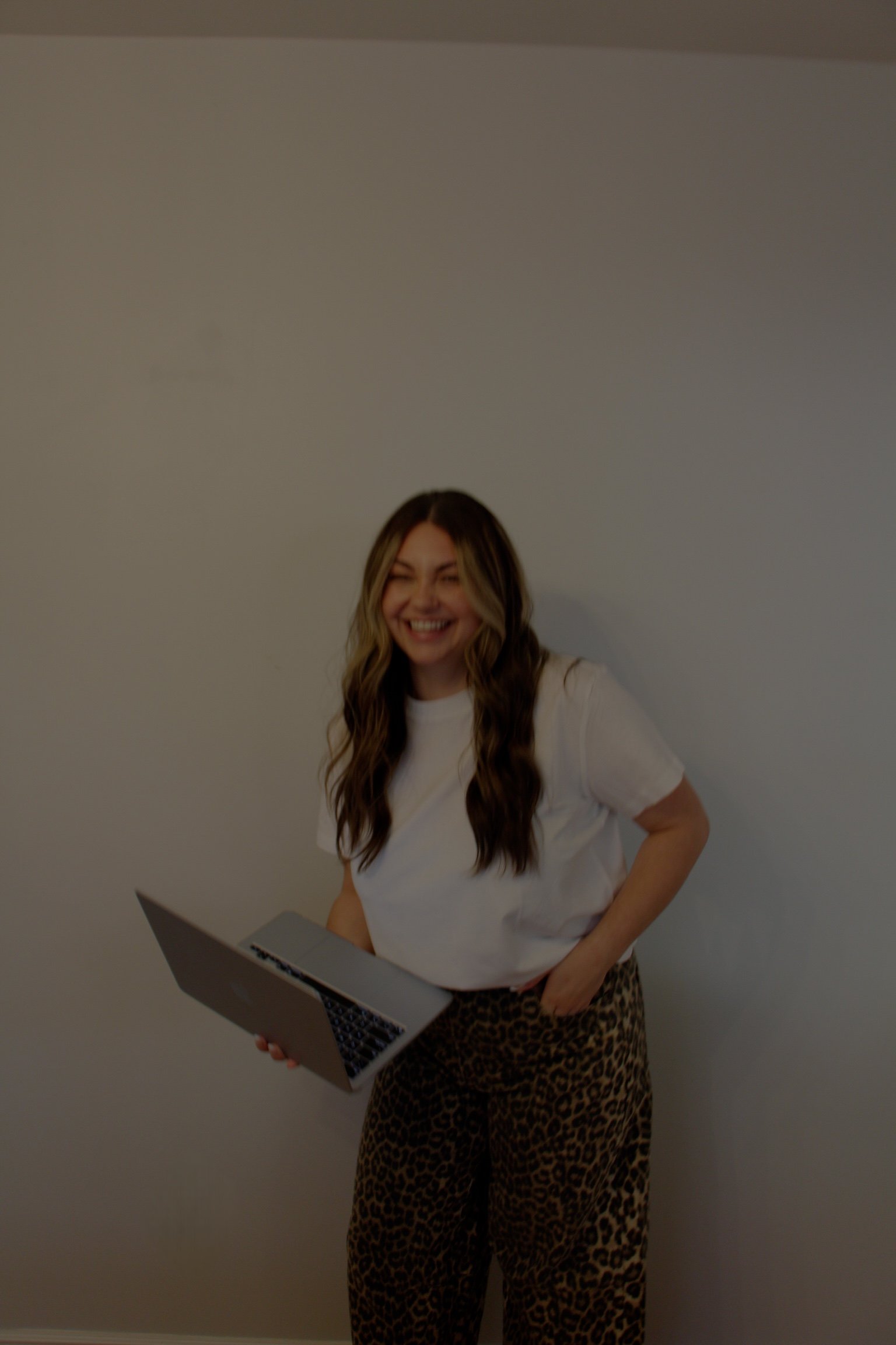 Woman with long wavy hair smiling, wearing a white t-shirt and leopard print pants, holding a laptop, standing against a plain wall.