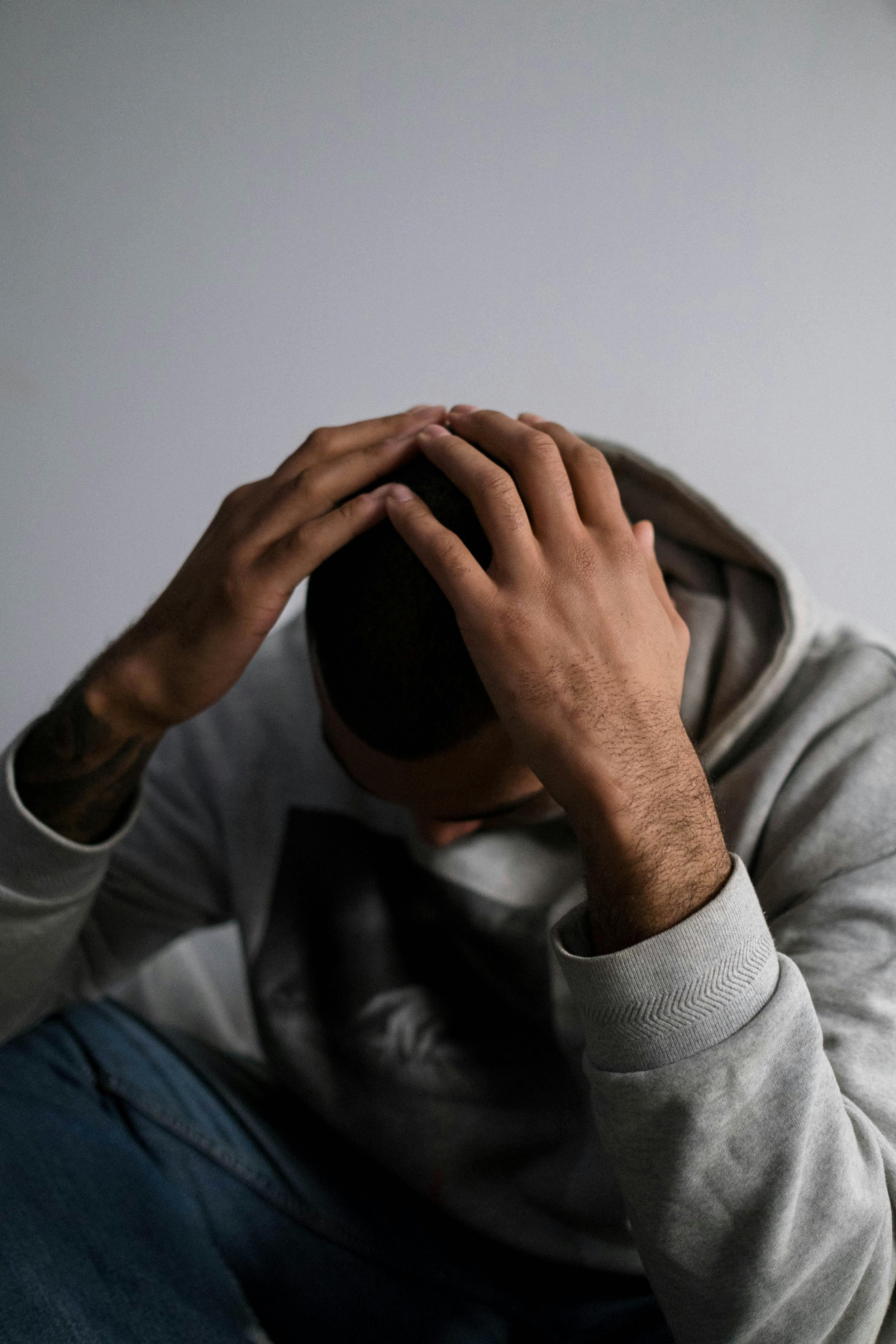 A person with dark hair, wearing a gray hoodie, sitting with their head bowed and their hands clasped on top of their head, against a plain light-colored wall.