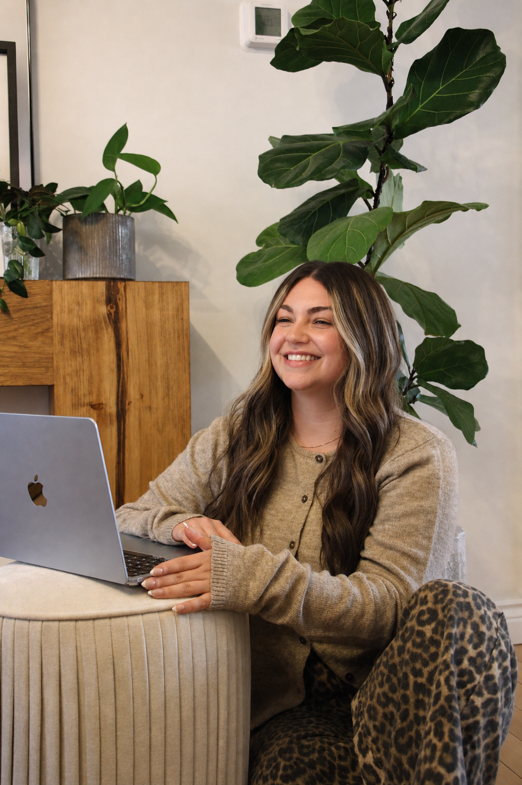 A young woman with long wavy hair smiling while sitting at a table with a laptop in a cozy, plant-filled room.