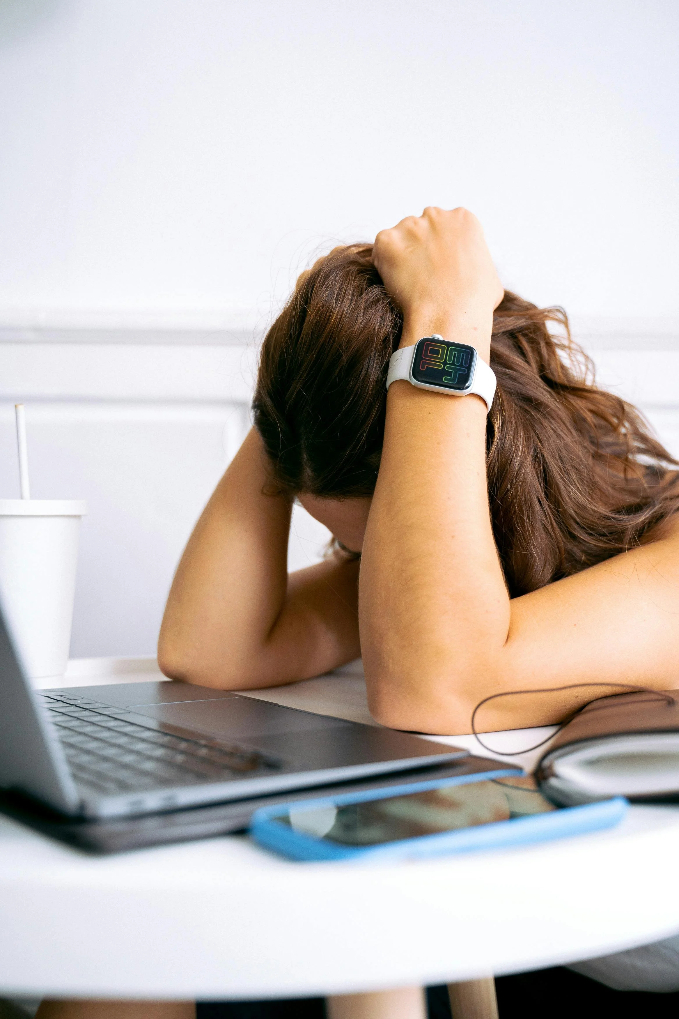 Woman with long brown hair resting her head on her hand, sitting at a white desk with a laptop, wearing a smartwatch on her wrist, in a bright room.