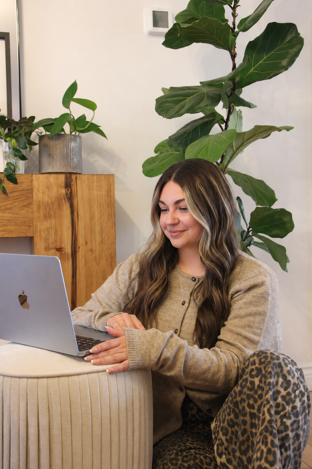 A woman with long wavy hair working on a silver MacBook laptop, sitting on a beige, pleated round stool in a cozy space with plants and wooden furniture.