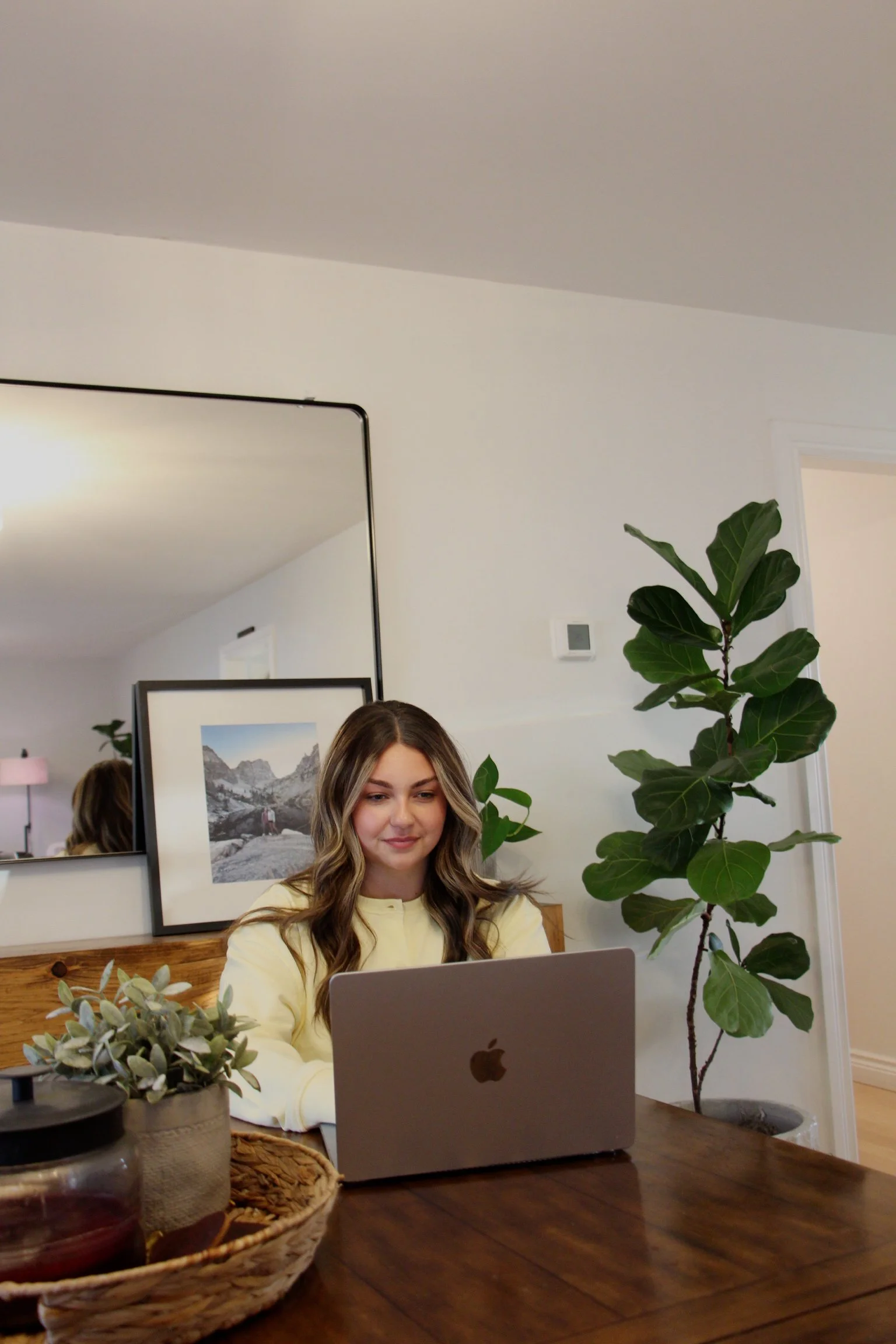 A woman with long wavy hair working on a laptop at a wooden table, surrounded by green plants and decor, with a mirror and framed pictures on the wall behind her.