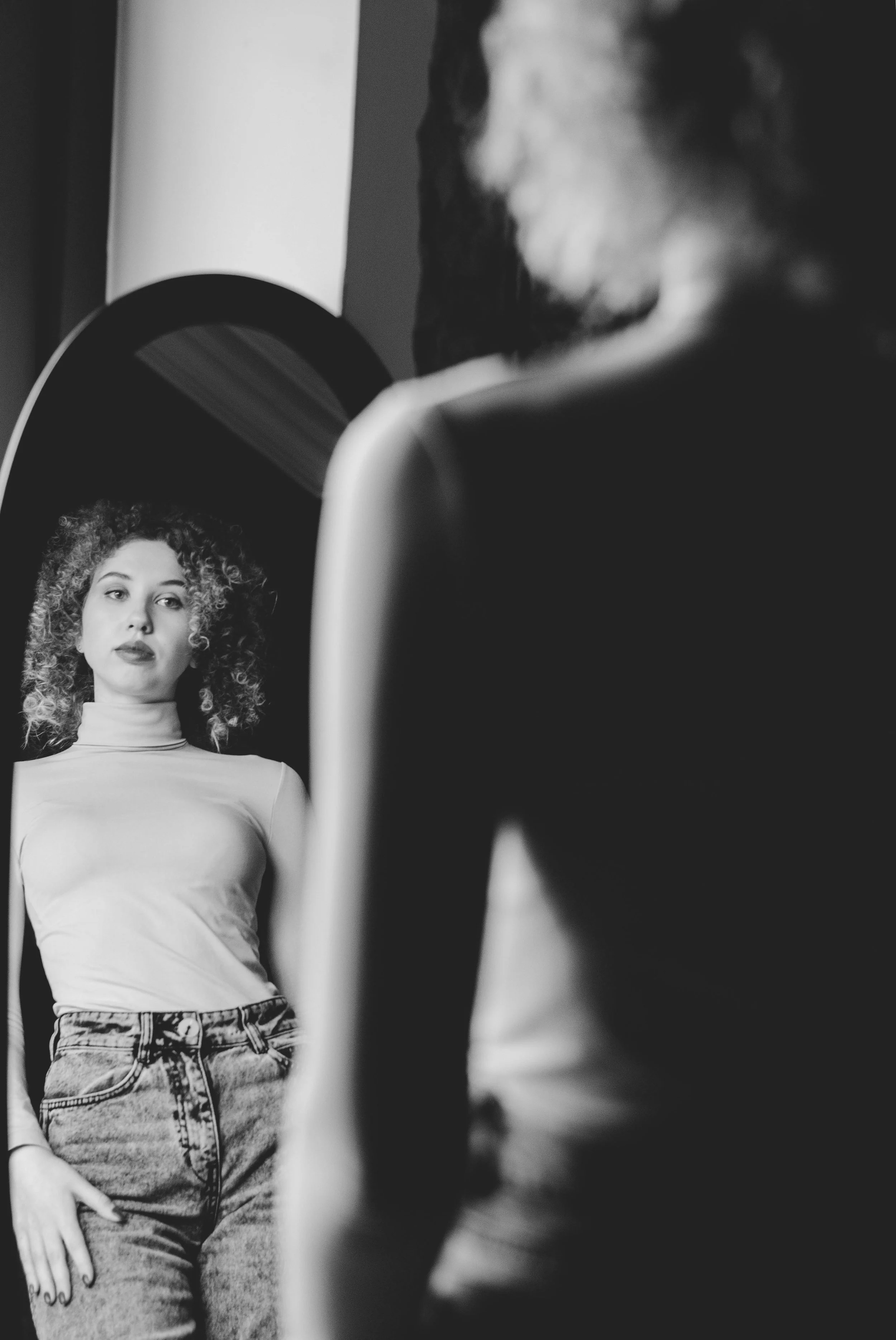 Black and white photo of a young woman with curly hair, wearing a turtleneck and jeans, looking at her reflection in a mirror.