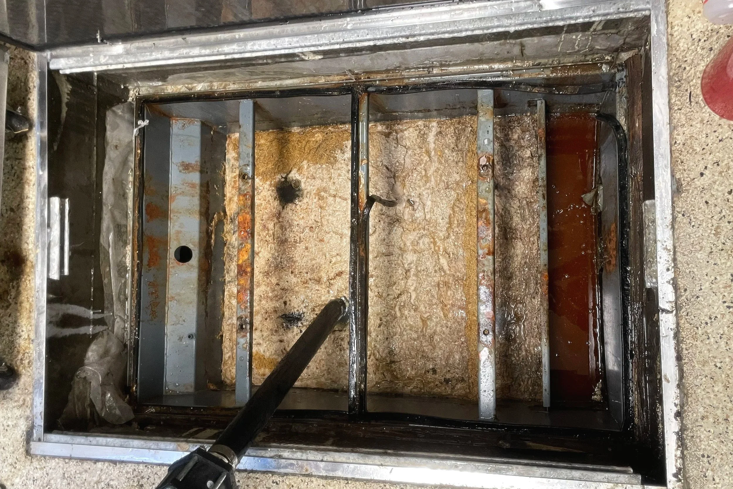 Dirty grease trap with rusty metal bars and black buildup inside a rectangular metal frame.