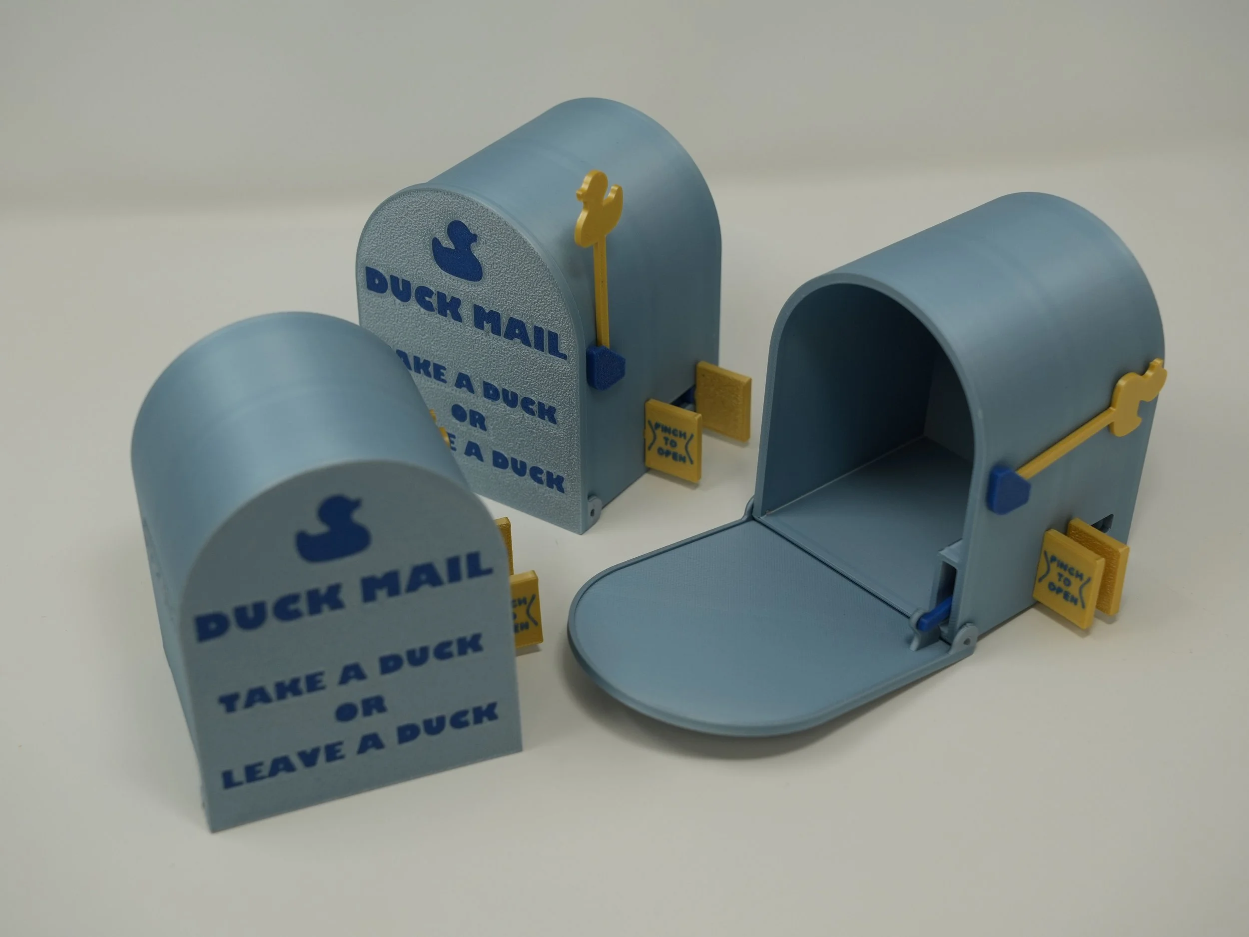Three small blue toy mailboxes with yellow and blue openers, one with a yellow flag, and one open. Each mailbox has the words "Duck Mail" and "Take a Duck or Leave a Duck" written on them.