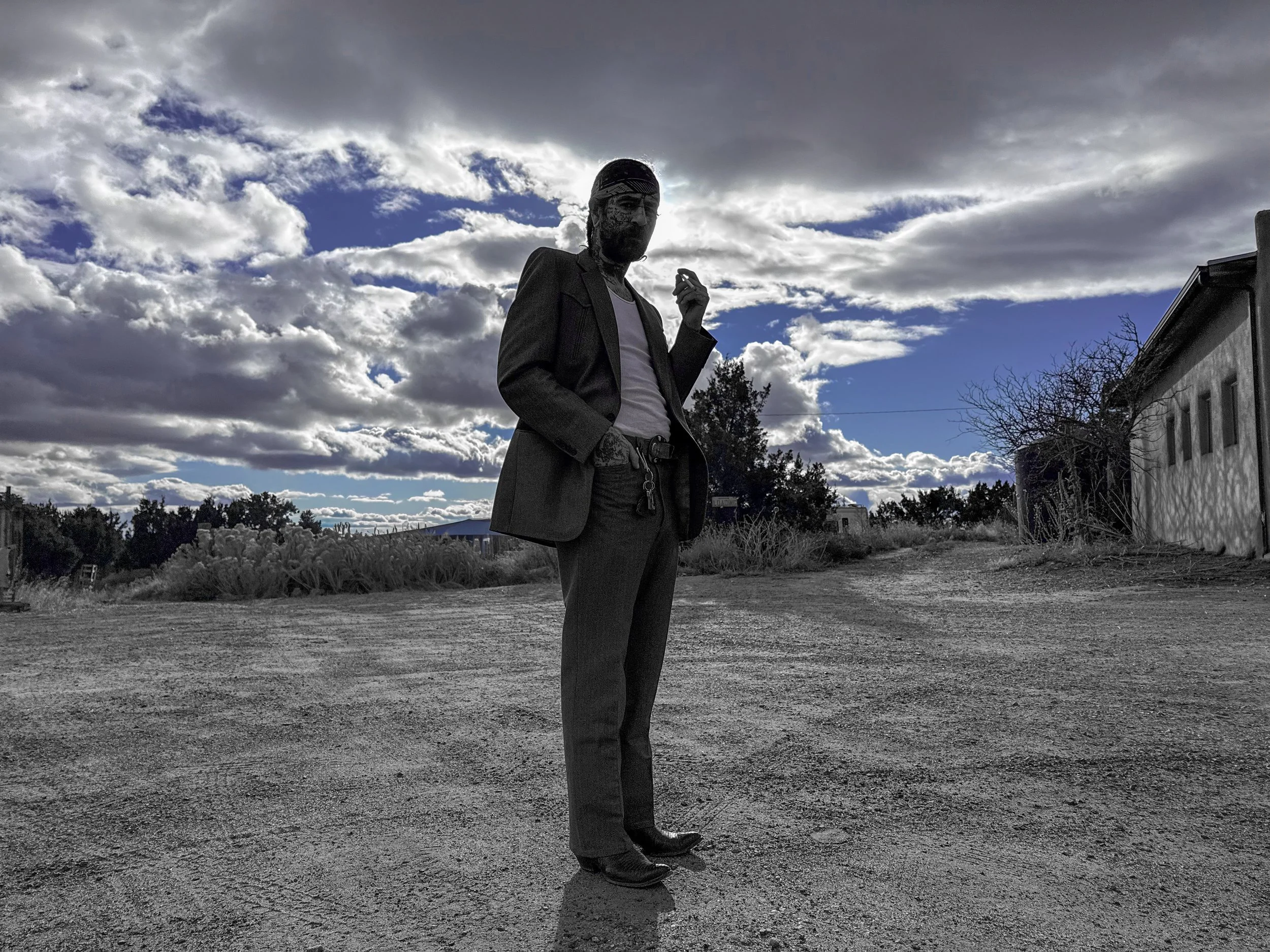 A man dressed in a suit with a white shirt and sunglasses is standing outdoors on a dirt path, smoking a cigarette, with a cloudy sky in the background.