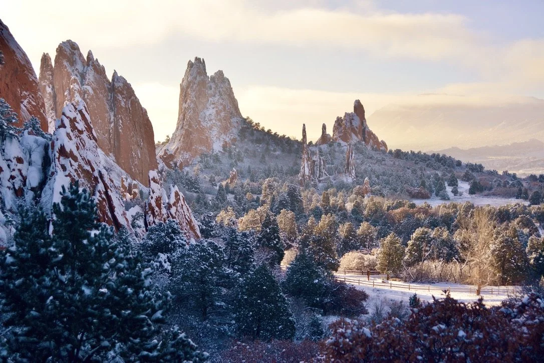 Snow-covered red rock formations and pine trees in a mountainous landscape during winter with a partly cloudy sky.