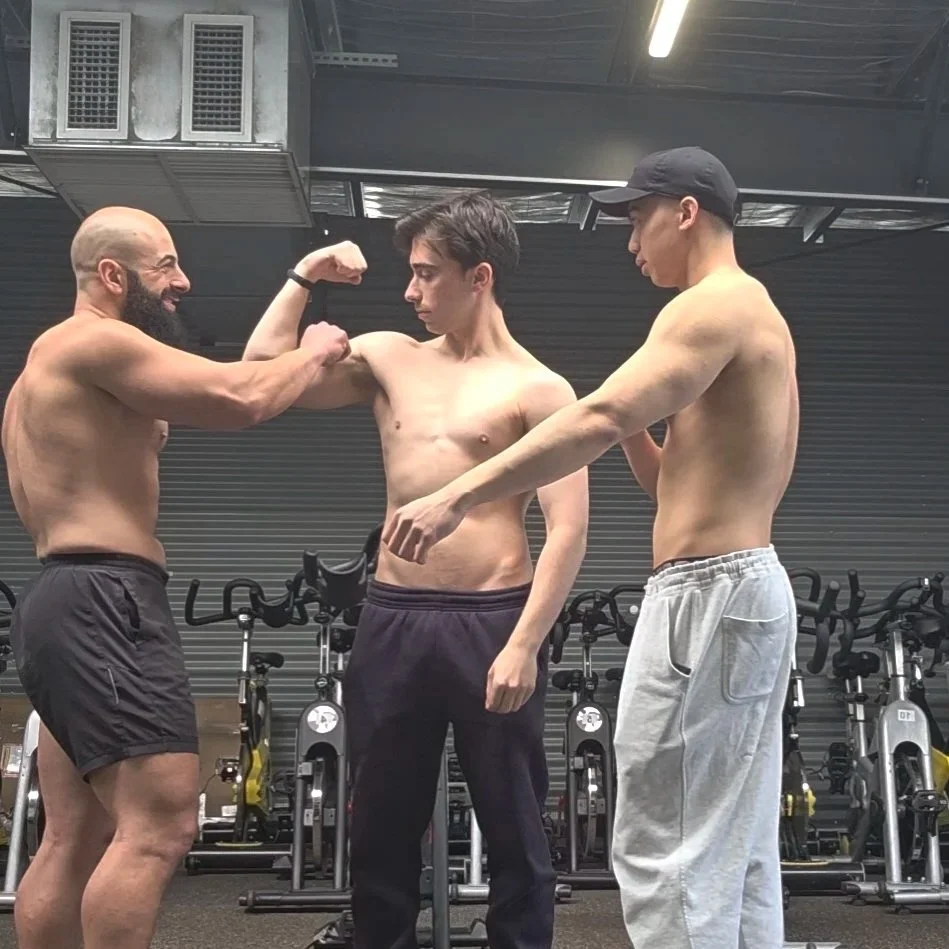 Two shirtless men and one shirted man at the gym, with one man flexing his arm while the other two observe, surrounded by exercise bikes.