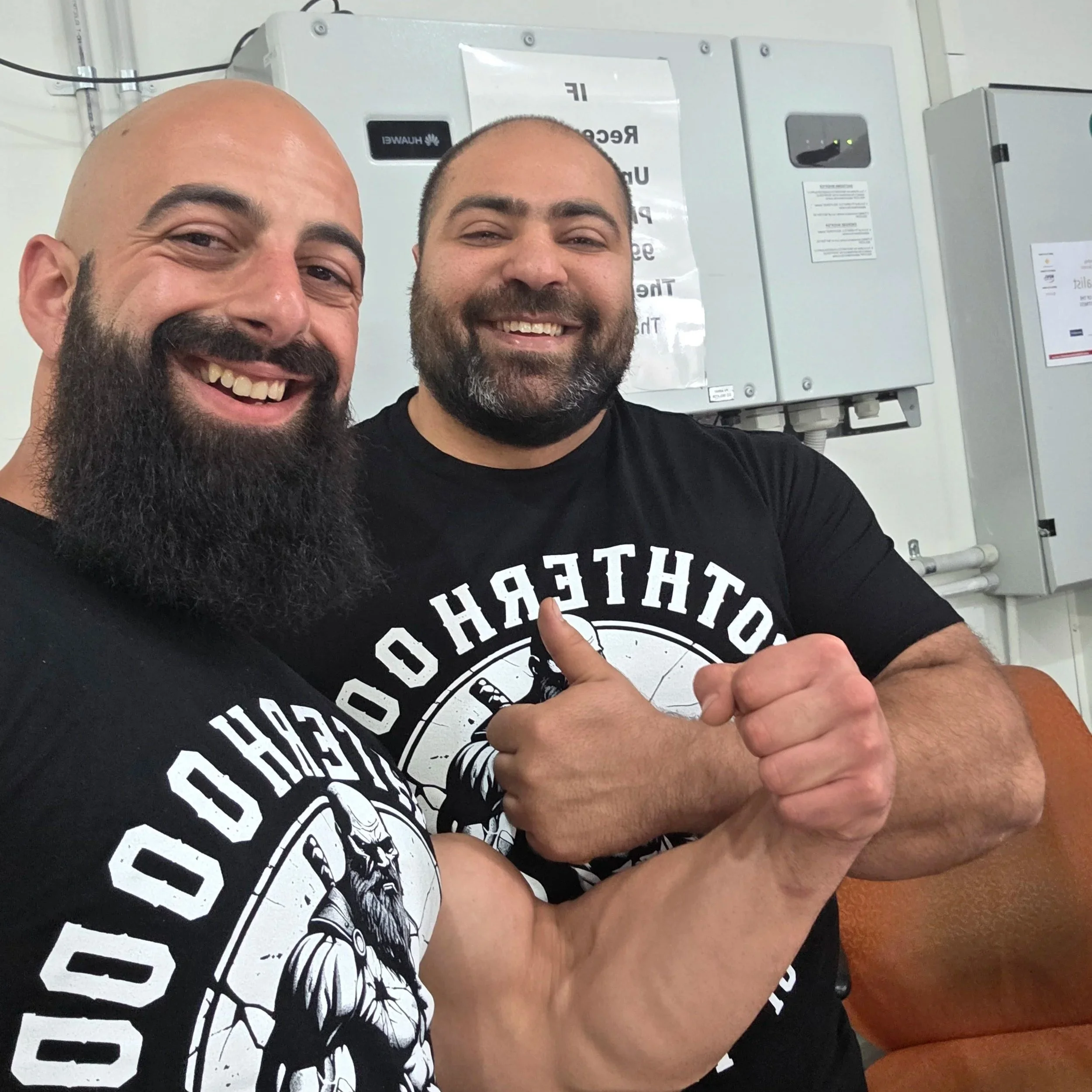 Two men smiling and showing off their biceps, wearing matching black T-shirts with a graphic and text, standing in front of electrical panels.