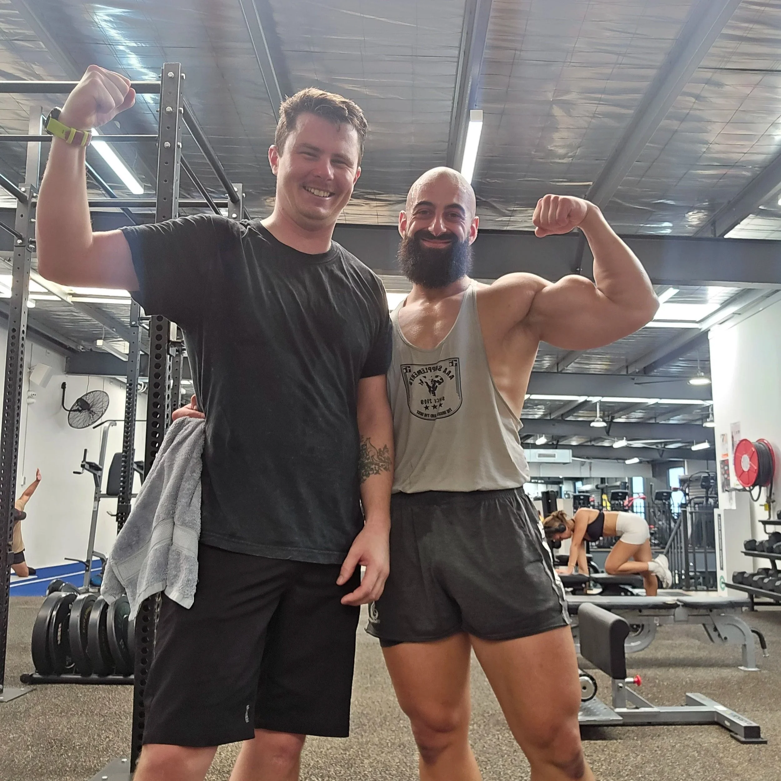 Two men at the gym are flexing their muscles and smiling at the camera, with weightlifting equipment and a woman working out in the background.