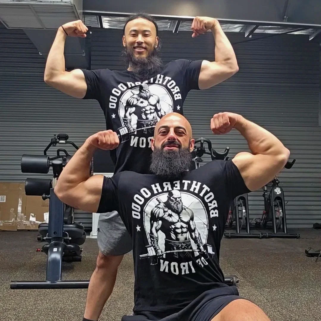 Two muscular men flexing their biceps at a gym, both wearing black t-shirts with a muscular warrior graphic and the text 'BETHESDAOOD of IRON'. One man is standing behind the other, who is seated, both showing their arm muscles and smiling.