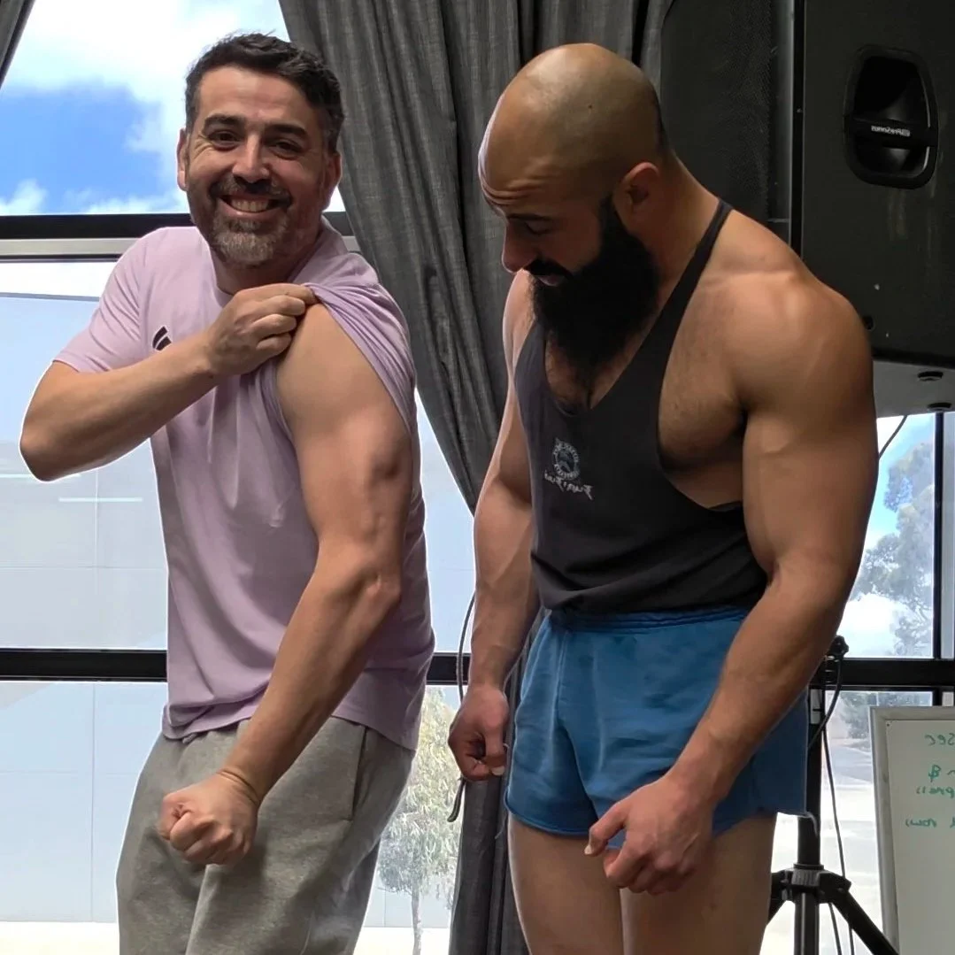 Two men with muscular arms, one showing his bicep to the camera smiling, the other looking down at his own arm, in casual clothes indoors near a window.