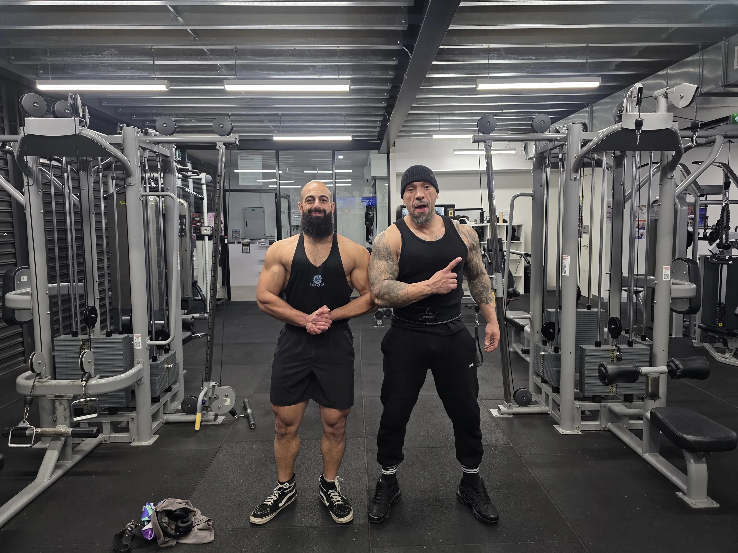 Two muscular men in black workout clothes posing in a gym with exercise equipment in the background.
