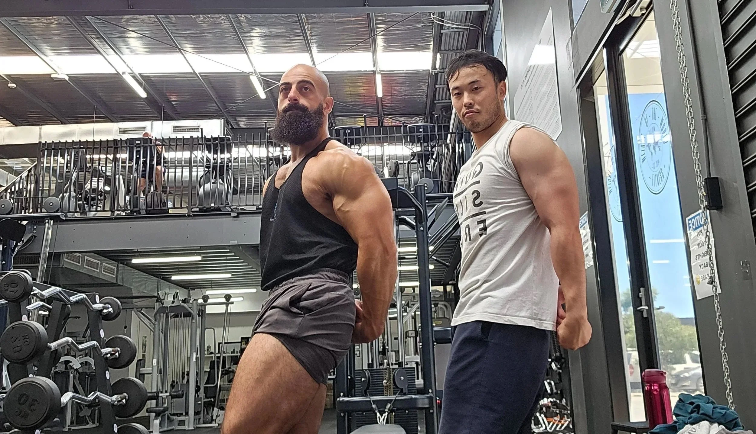 Two muscular men posing in a gym, with weight equipment and cardio machines in the background.