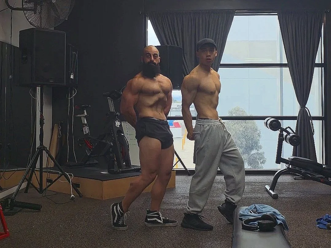 Two shirtless men with muscular physiques pose in a gym, one wearing black shorts and the other gray sweatpants. They stand in front of gym equipment and large windows with dark curtains.