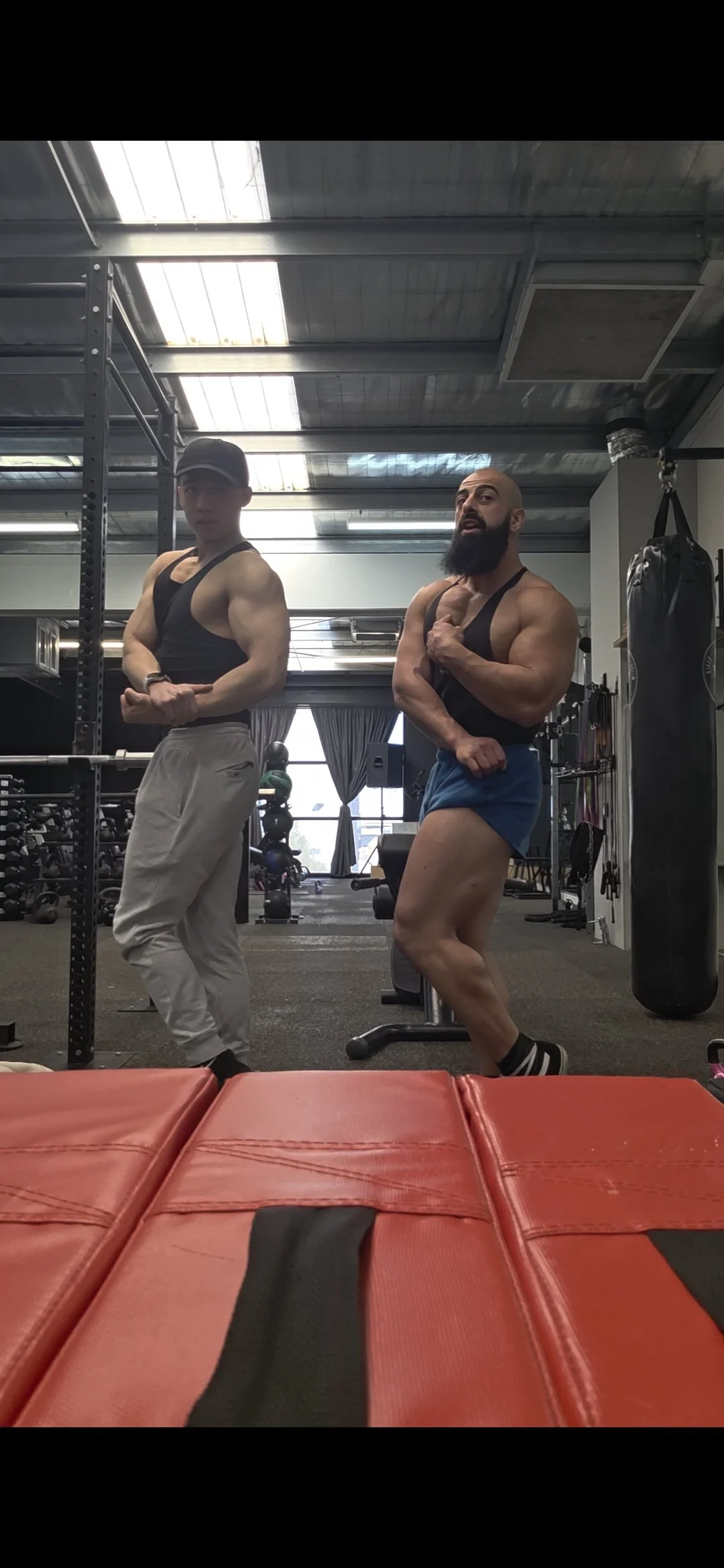 Two muscular men posing in a gym, one with a beard and one wearing a cap, with gym equipment in the background.