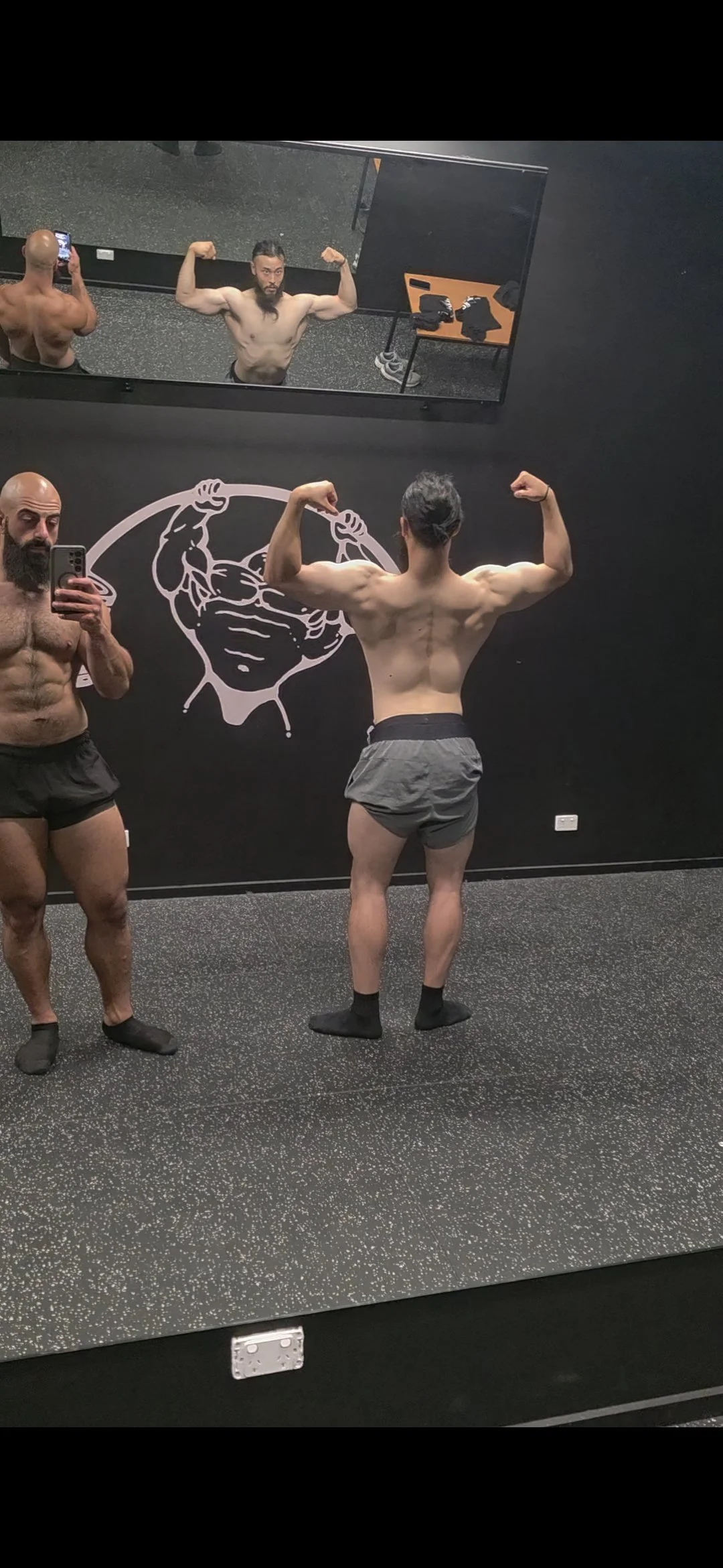 Two men with muscular physiques, shirtless, taking a mirror selfie in a gym. One man is facing the mirror, flexing his biceps, and the other man, with a beard, is holding a phone and standing beside him. The gym has a black wall with a muscular super