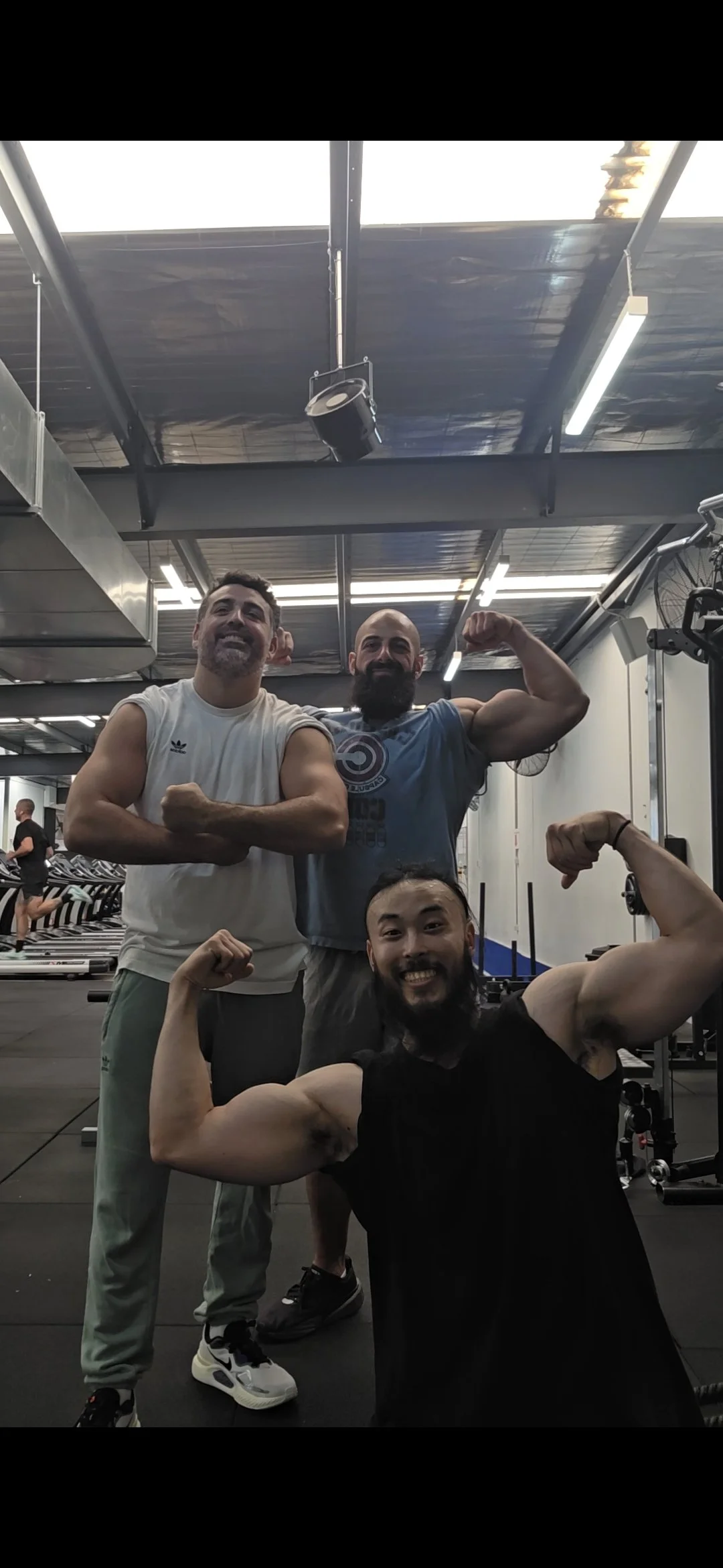 Three men in a gym posing and flexing their muscles, with cardio machines and gym equipment in the background.