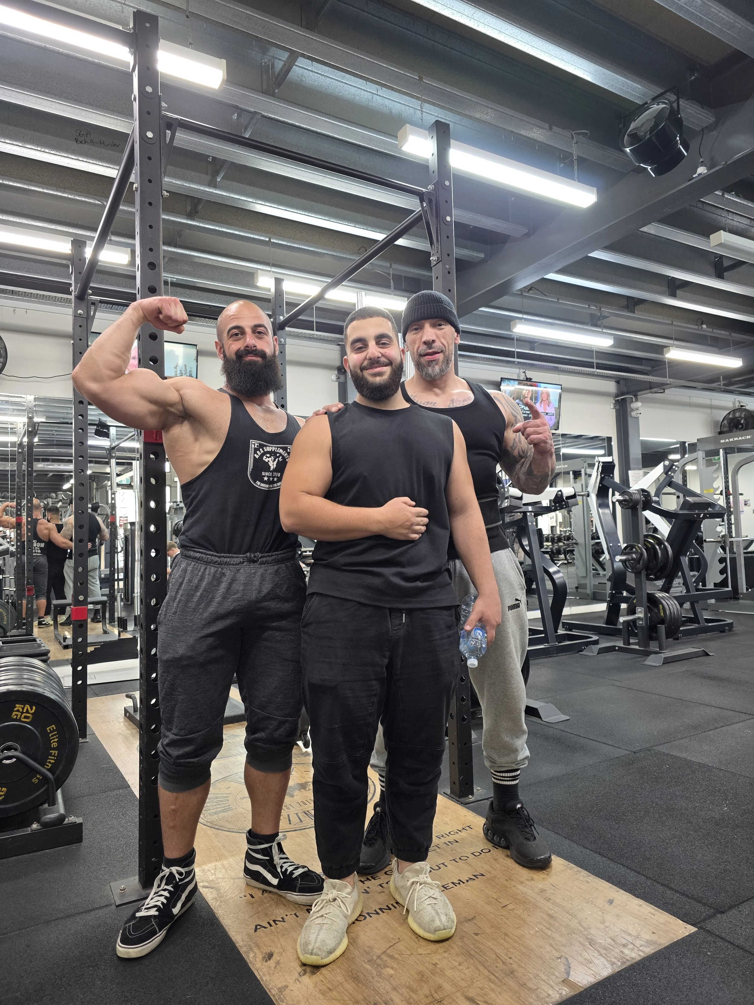 Three muscular men posing in a gym. The man on the left is flexing his bicep, the man in the middle is smiling with one hand on his stomach and the other hanging down, and the man on the right is giving a thumbs-up. The gym has weightlifting equipmen