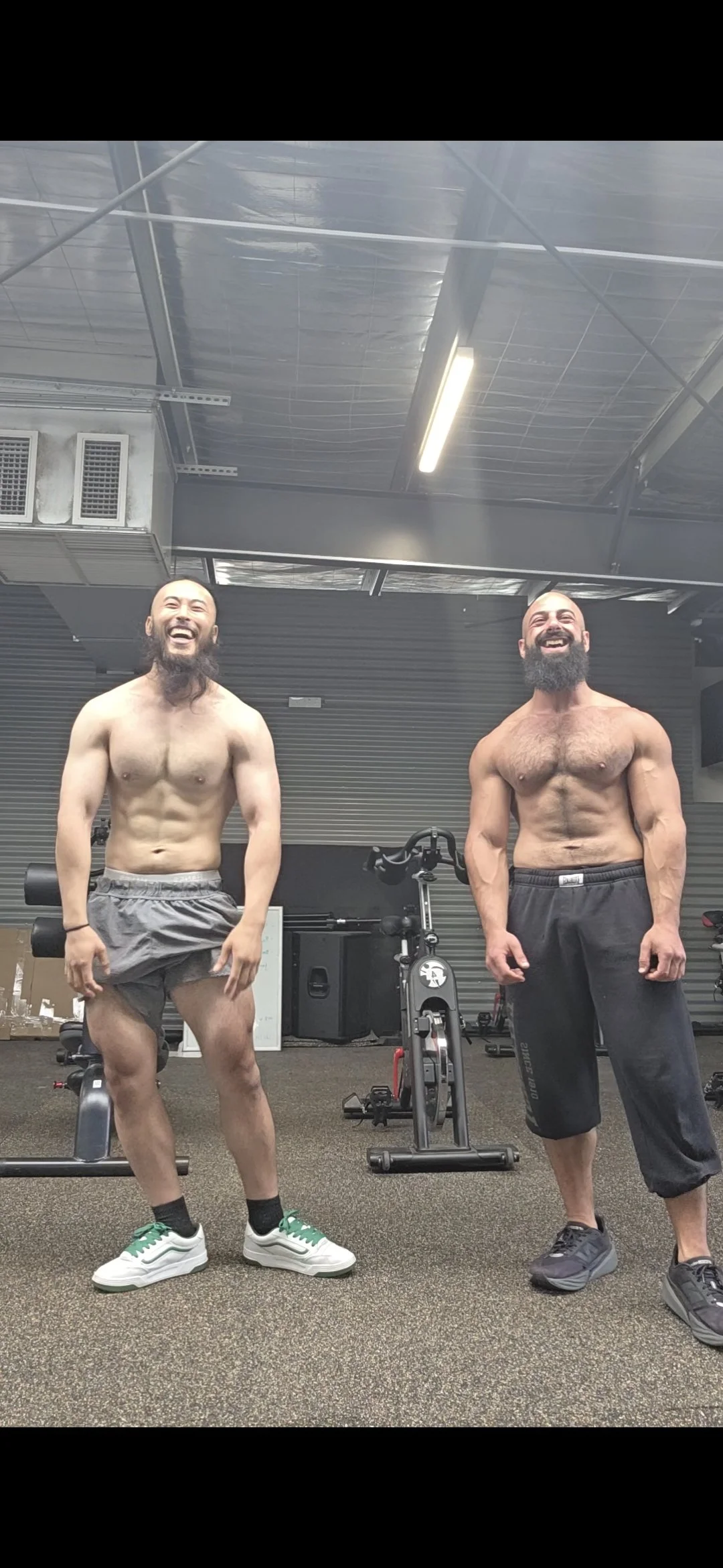 Two shirtless men with beards and muscular build, standing in a gym, smiling and laughing. The man on the left is wearing gray shorts and white sneakers, while the man on the right is wearing black shorts and black sneakers. Gym equipment is visible 