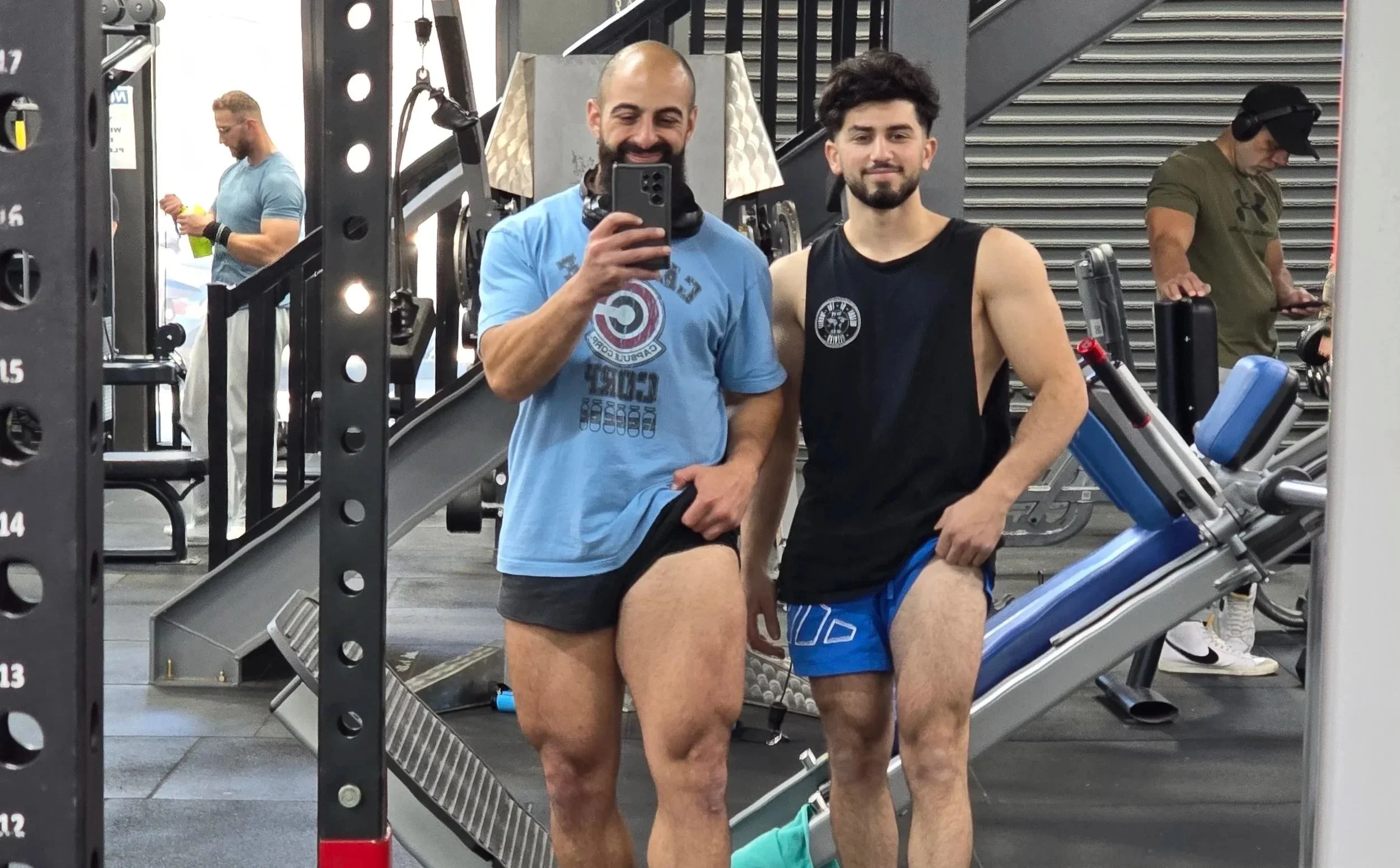 Two men in a gym taking a selfie in front of mirror, one wearing a light blue shirt and black shorts, the other in a black sleeveless shirt and blue shorts.