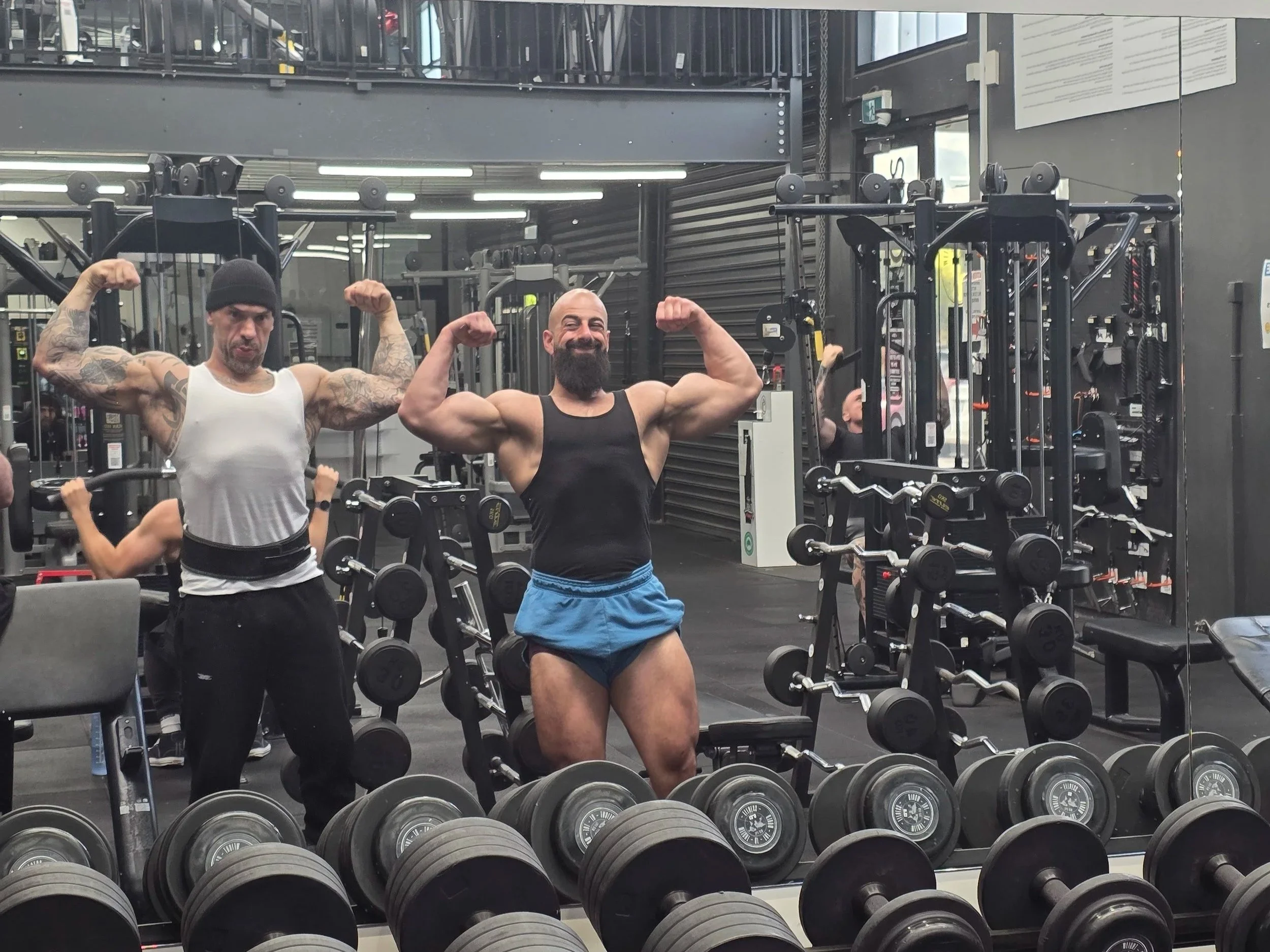 Two muscular men flexing their biceps and posing in a gym with weightlifting equipment and dumbbells in front of a mirror.