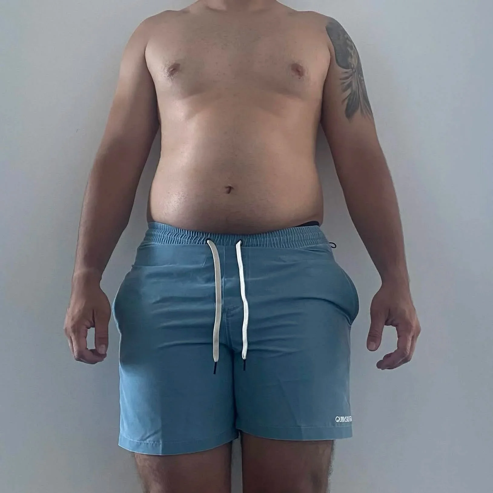 A shirtless man with athletic build and a tattoo on his left shoulder, standing against a plain wall, wearing blue athletic shorts with a white drawstring.