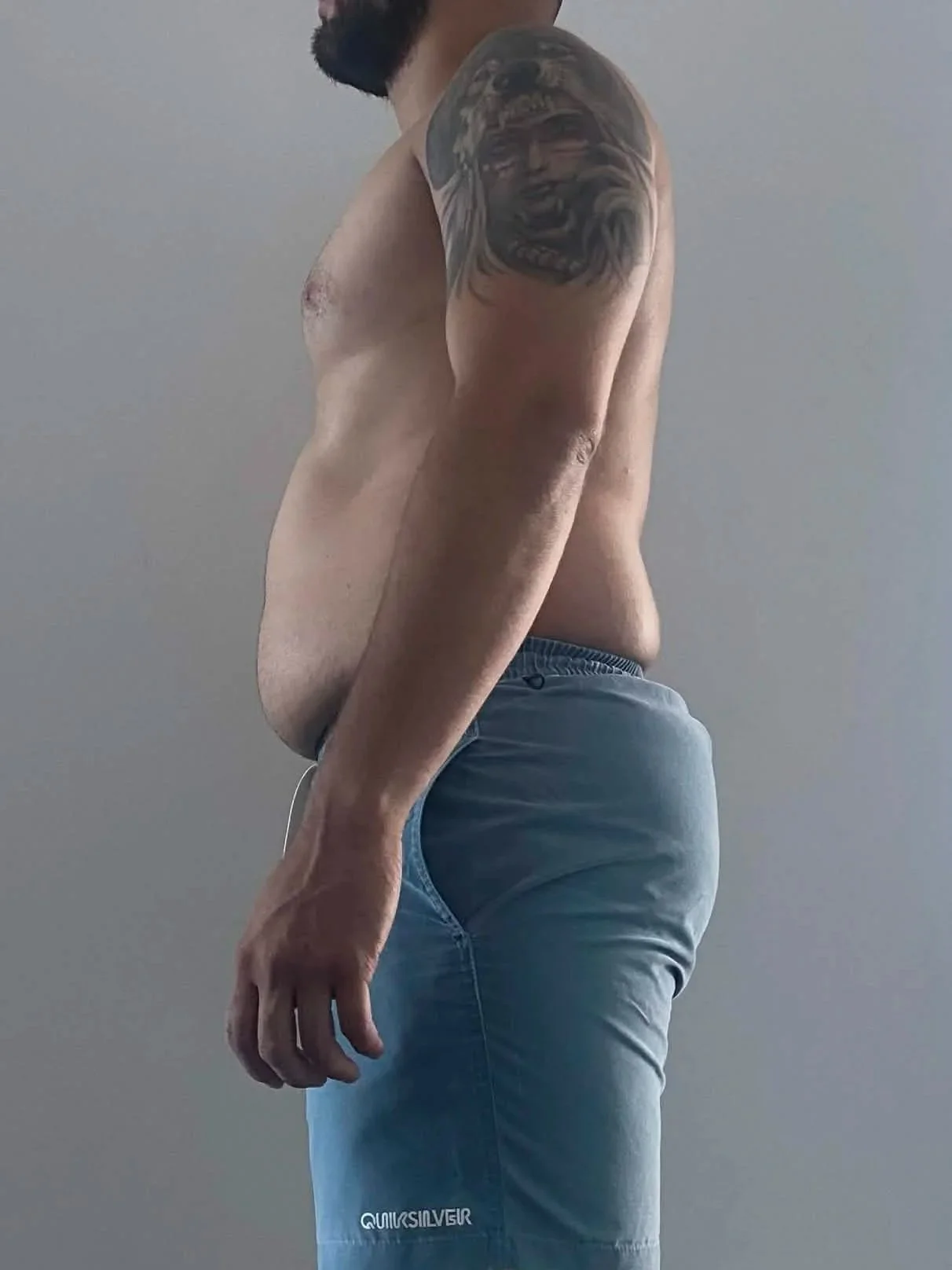 Side view of a shirtless man with a tattoo on his left arm, wearing blue shorts, standing against a plain background.