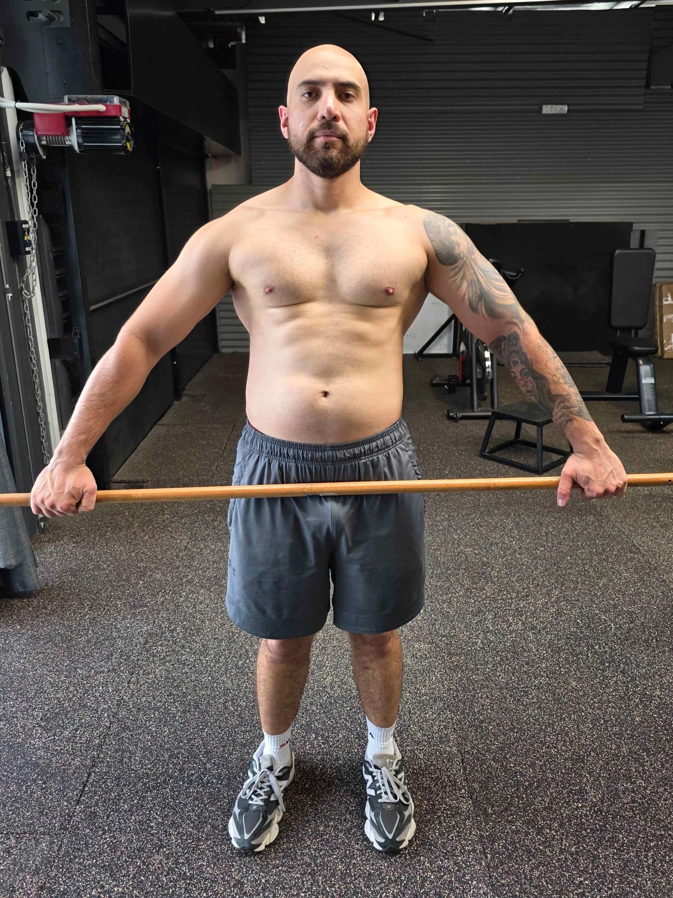 A shirtless man with a beard and tattoos holding a wooden stick in a gym.