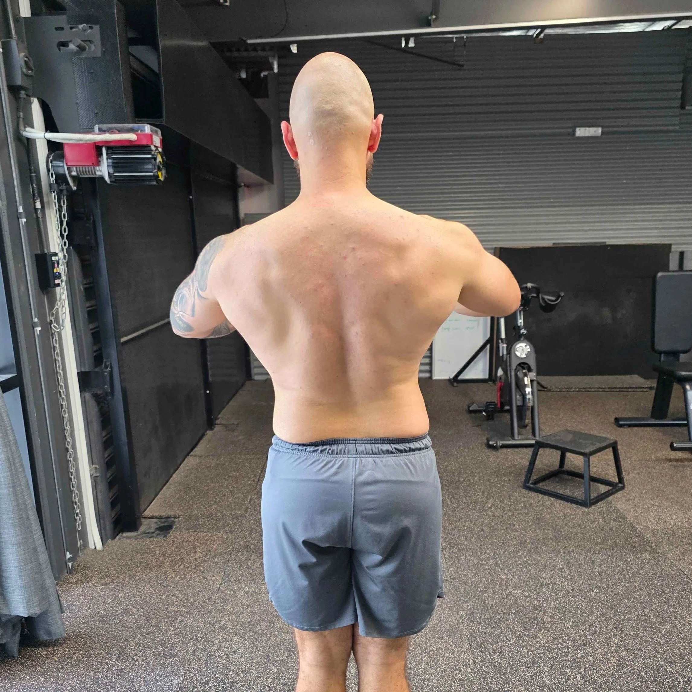 A muscular, shirtless man with a shaved head and a tattoo on his left arm, standing in a gym and facing away from the camera.
