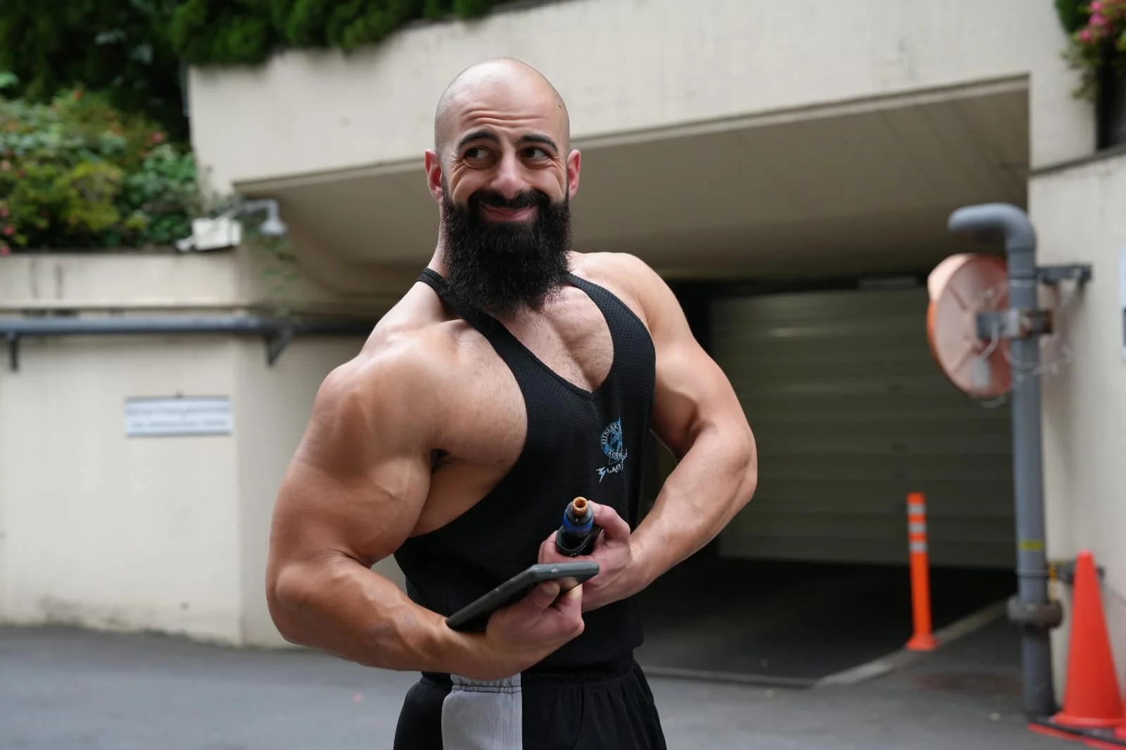 A muscular man with a beard, wearing a black tank top, holding a cellphone and a bottle, standing outdoors near a parking garage.