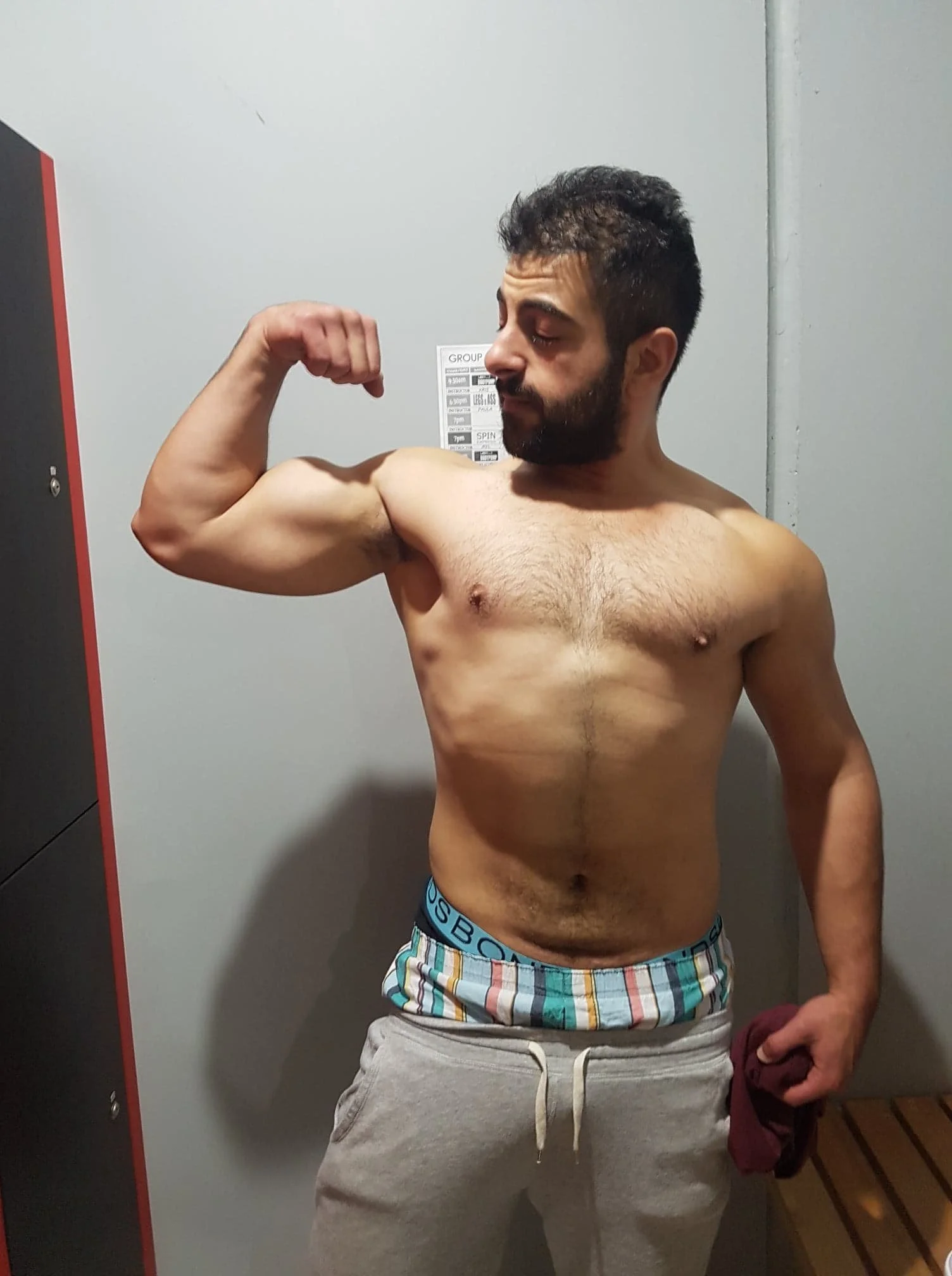 A shirtless man with a beard and short hair flexing his bicep while looking at it in a room with gray walls.