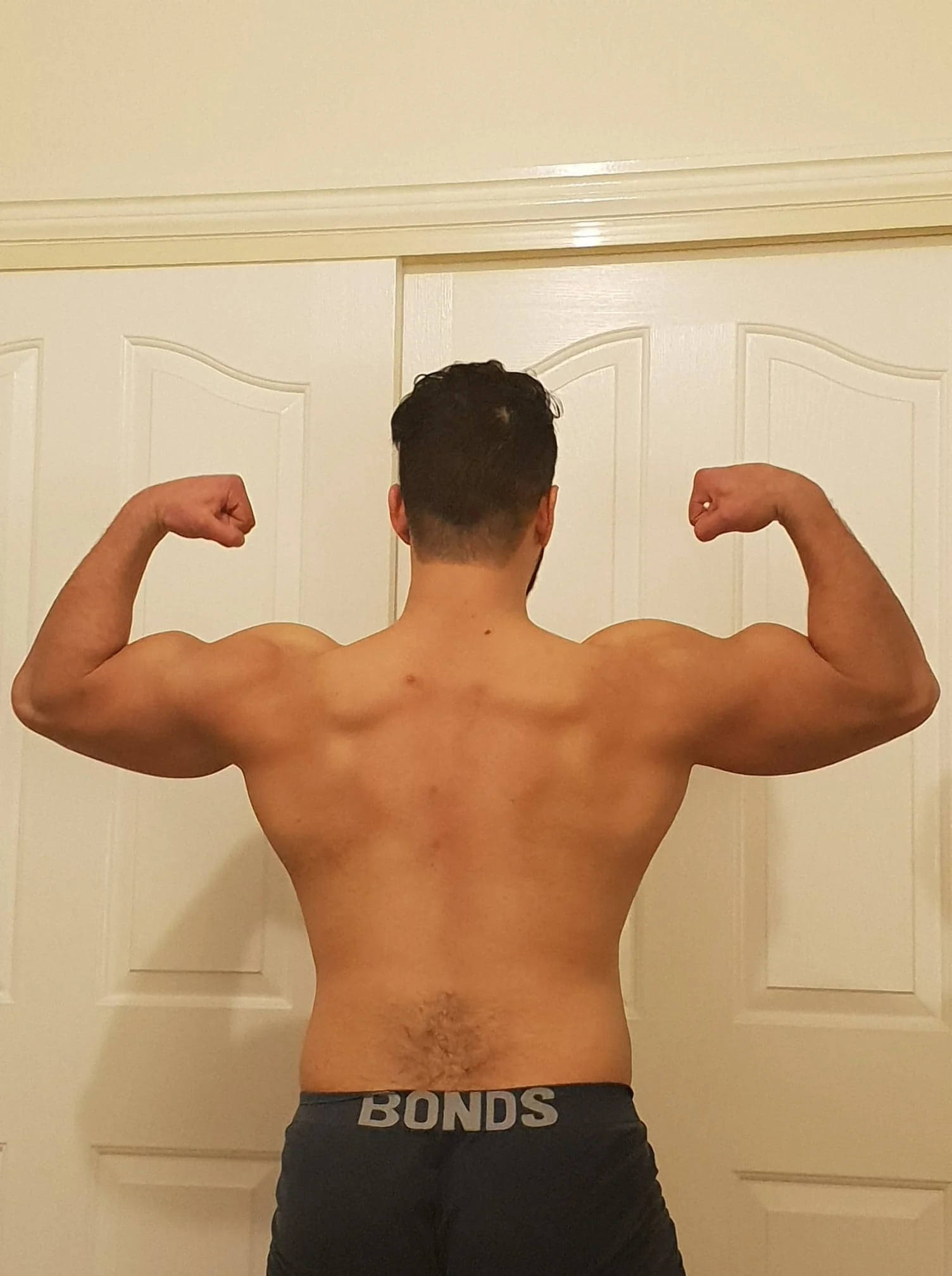 Back view of a muscular man flexing his biceps, wearing black shorts with 'BONDS' printed on the waistband, standing in front of a closed white door.