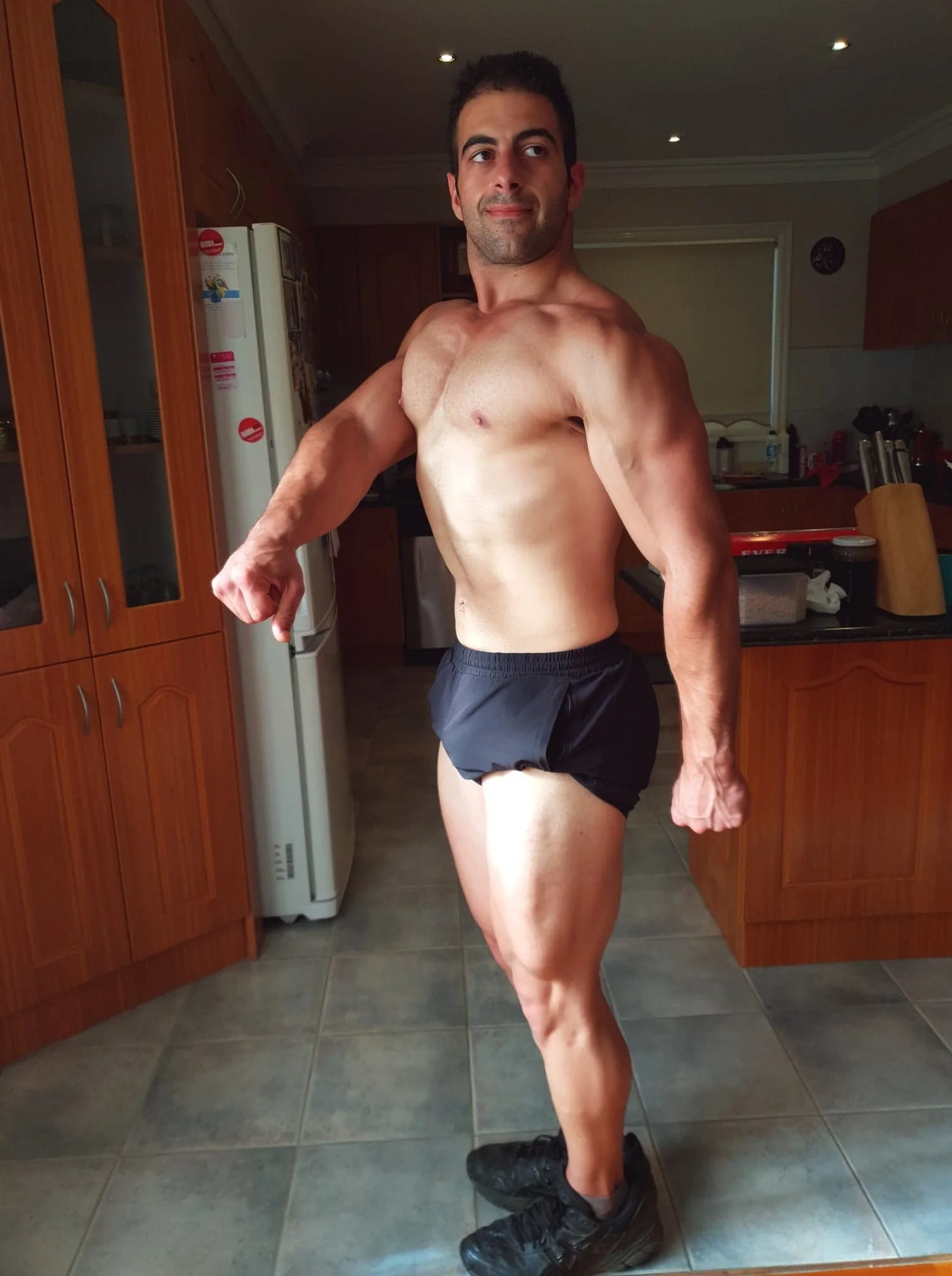 A muscular man standing in a kitchen, wearing black running shoes and black shorts, flexing his left arm while looking at the camera.