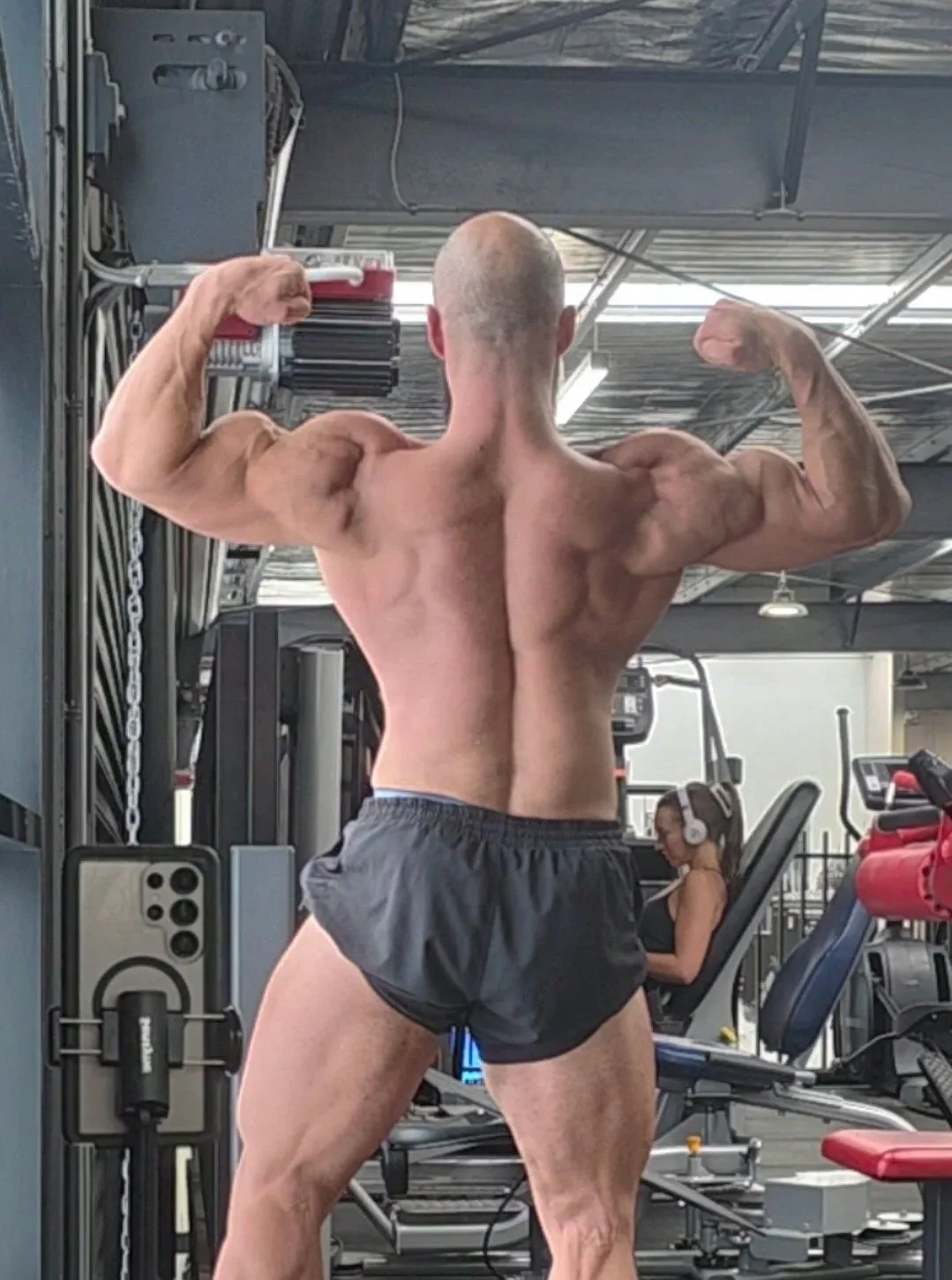 A muscular man flexes his back and arm muscles in a gym, wearing black shorts, with various workout equipment around him and a woman in headphones working out in the background.