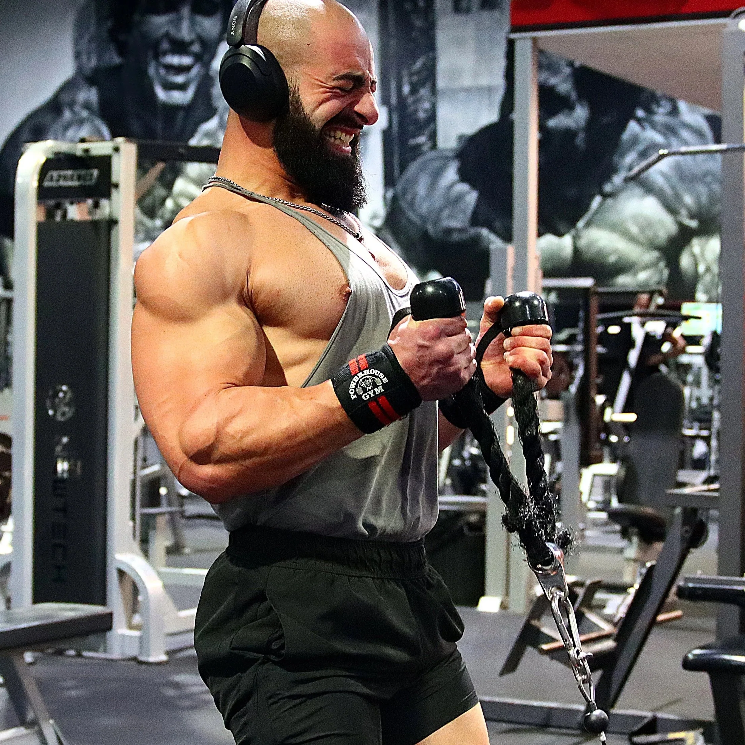 A muscular man with a beard and headphones working out with a tricep rope at a gym, smiling and showing his biceps.