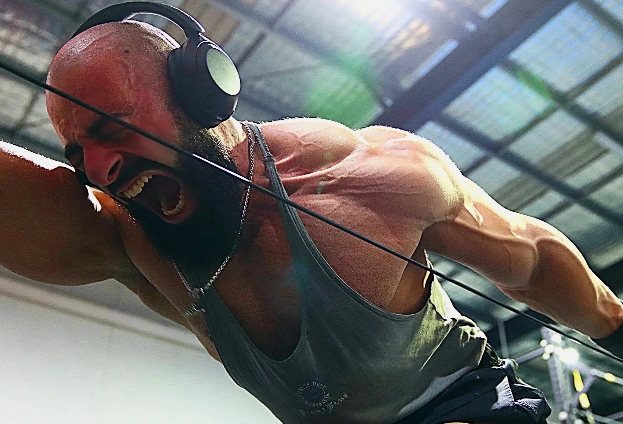 A muscular man with a beard wearing headphones is intensely working out, likely doing a pull-up or similar exercise, in a gym with metal ceiling structures and sunlight shining through.