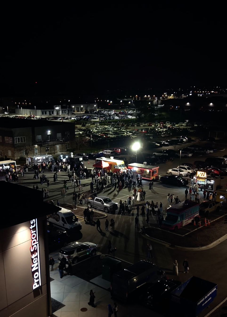 Nighttime scene of a parking lot outside a ProNet Sports store, filled with people attending an event with food trucks and tents, with additional parking lots and city lights in the background.