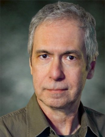 A middle-aged man with gray hair wearing a dark green collared shirt, looking directly at the camera with a neutral expression, against a dark blurred background.