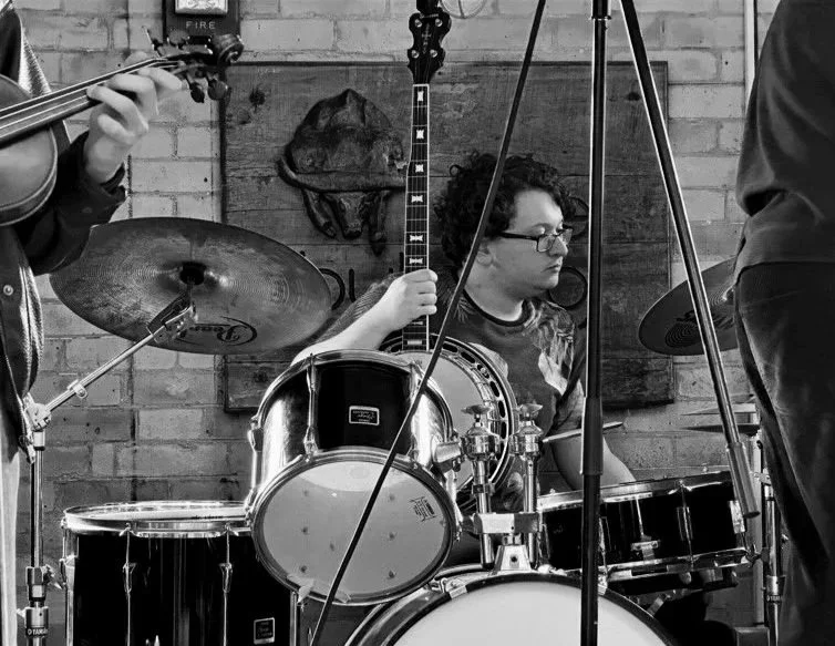 A young person with glasses playing drums in a band, with a violinist partly visible on the left and another musician on the right, against a brick wall background.