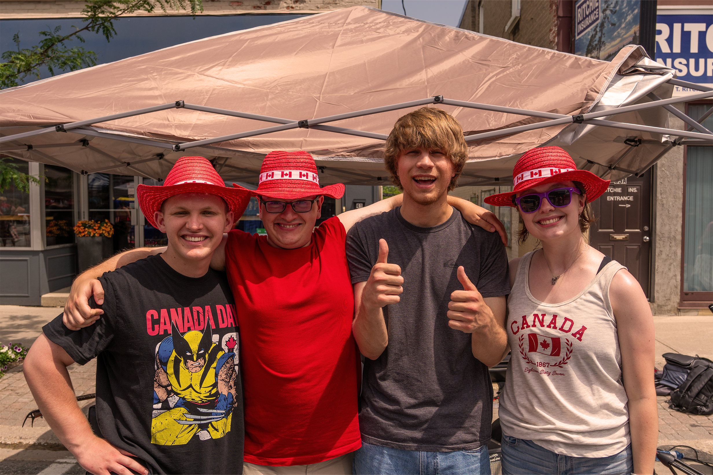 Four young people standing together under a beige canopy, smiling and wearing red hats with Canadian flags, with a storefront in the background.