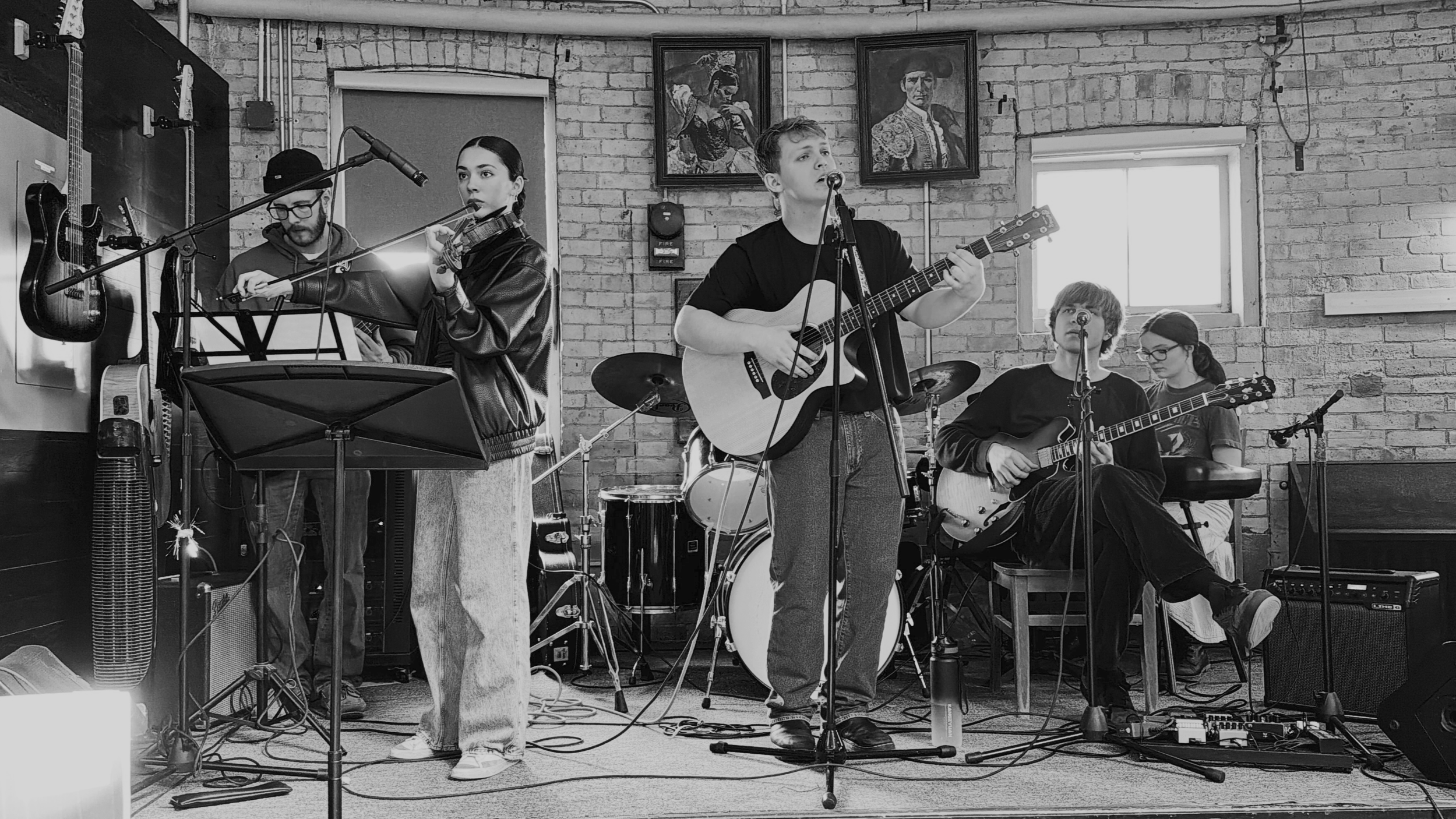 A group of five young musicians performing indoors against a brick wall. Two are playing guitars, one is playing drums, one is playing keyboard, and one is singing with a microphone. There are two framed paintings on the wall behind them and a window