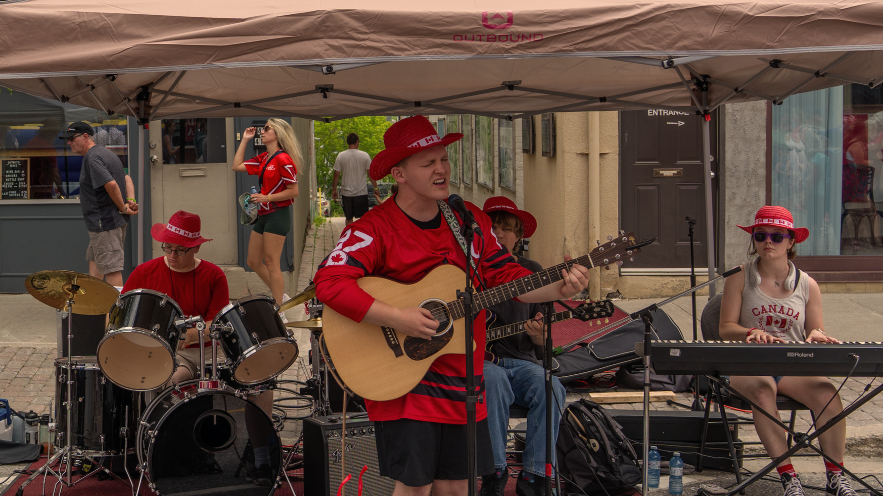 A group of children performing in a street band, with one playing drums, another singing and playing guitar, a third playing keyboard, all wearing red and Canadian-themed hats, under a beige canopy.