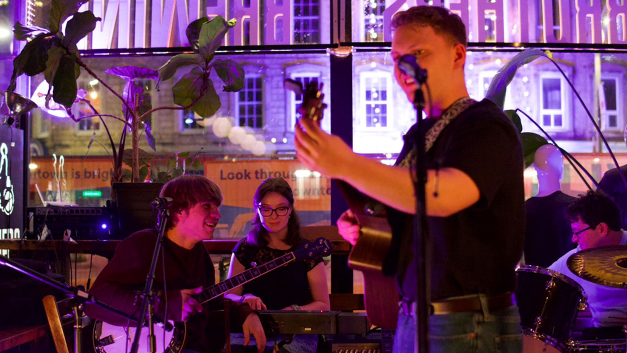 A group of people performing music in a lively indoor setting with colorful lighting, including a guitarist, a singer, and two women, one playing a keyboard and the other holding a guitar.