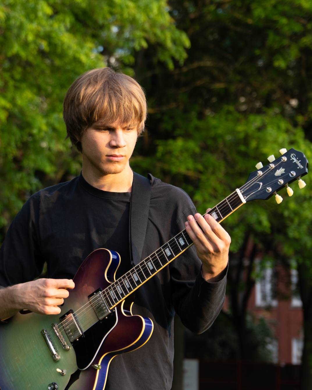 Young man playing an electric guitar outdoors with green trees in the background.
