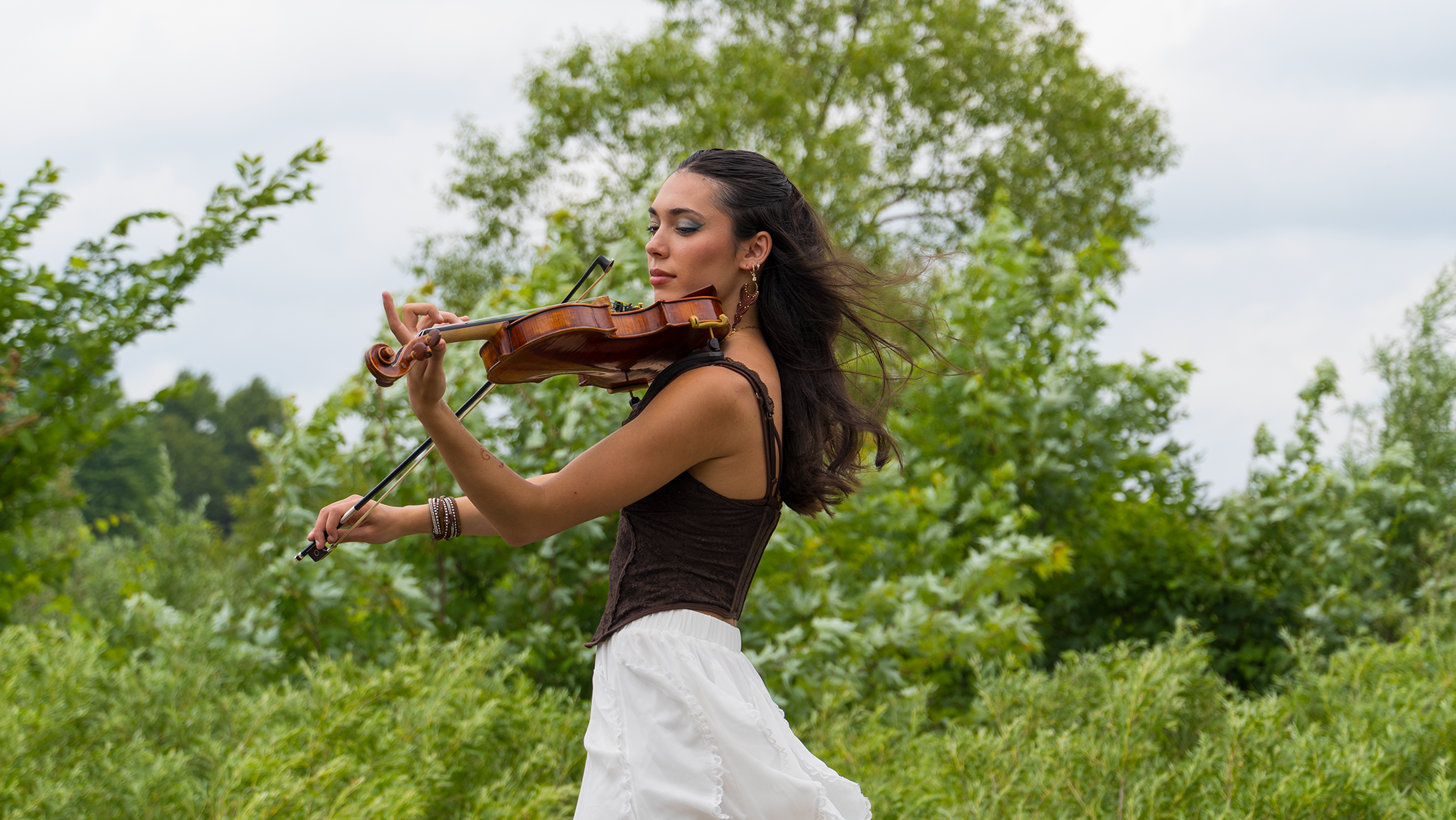 A woman playing a violin outdoors in a green park with trees in the background.