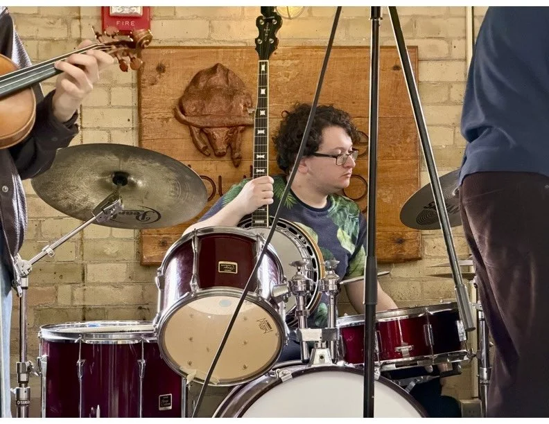 Young man with curly hair and glasses playing a drum set in a music rehearsal space with brick walls and wooden art on the background